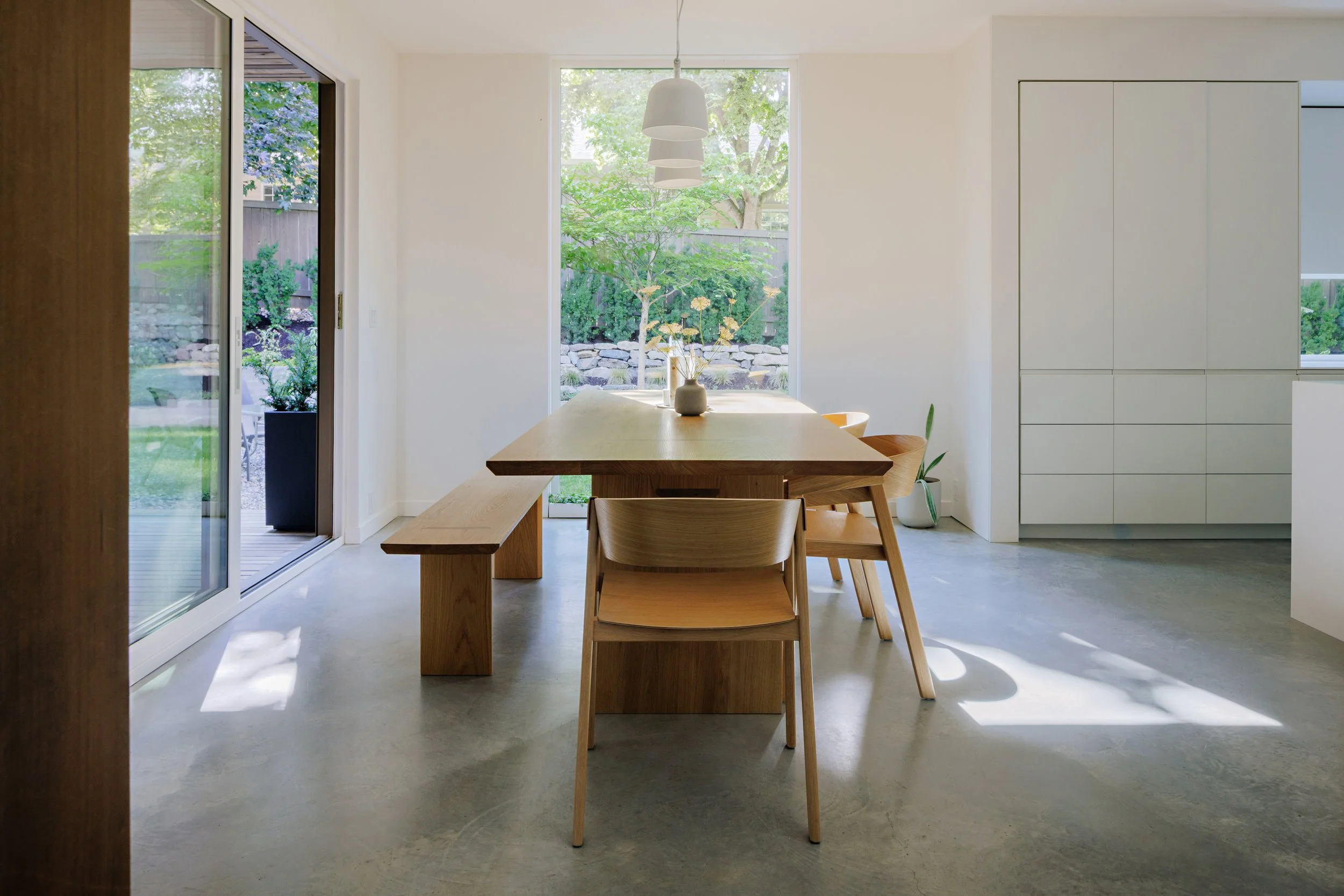 Modern dining area with a wooden table and matching chairs, large window revealing greenery outside, white walls, and minimalist decor.
