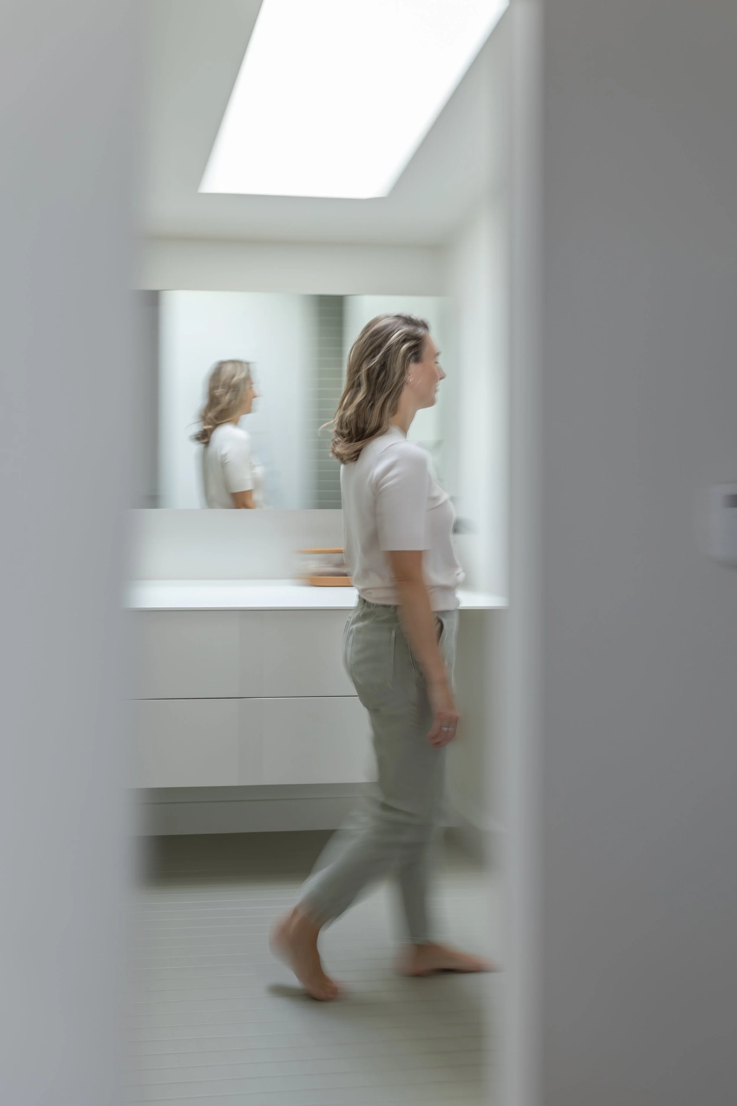 A woman with wavy blonde hair walking barefoot in a bright, modern room with a sloped skylight roof and a large mirror. She is dressed in a white blouse and light-colored pants with another reflection of her in the mirror.