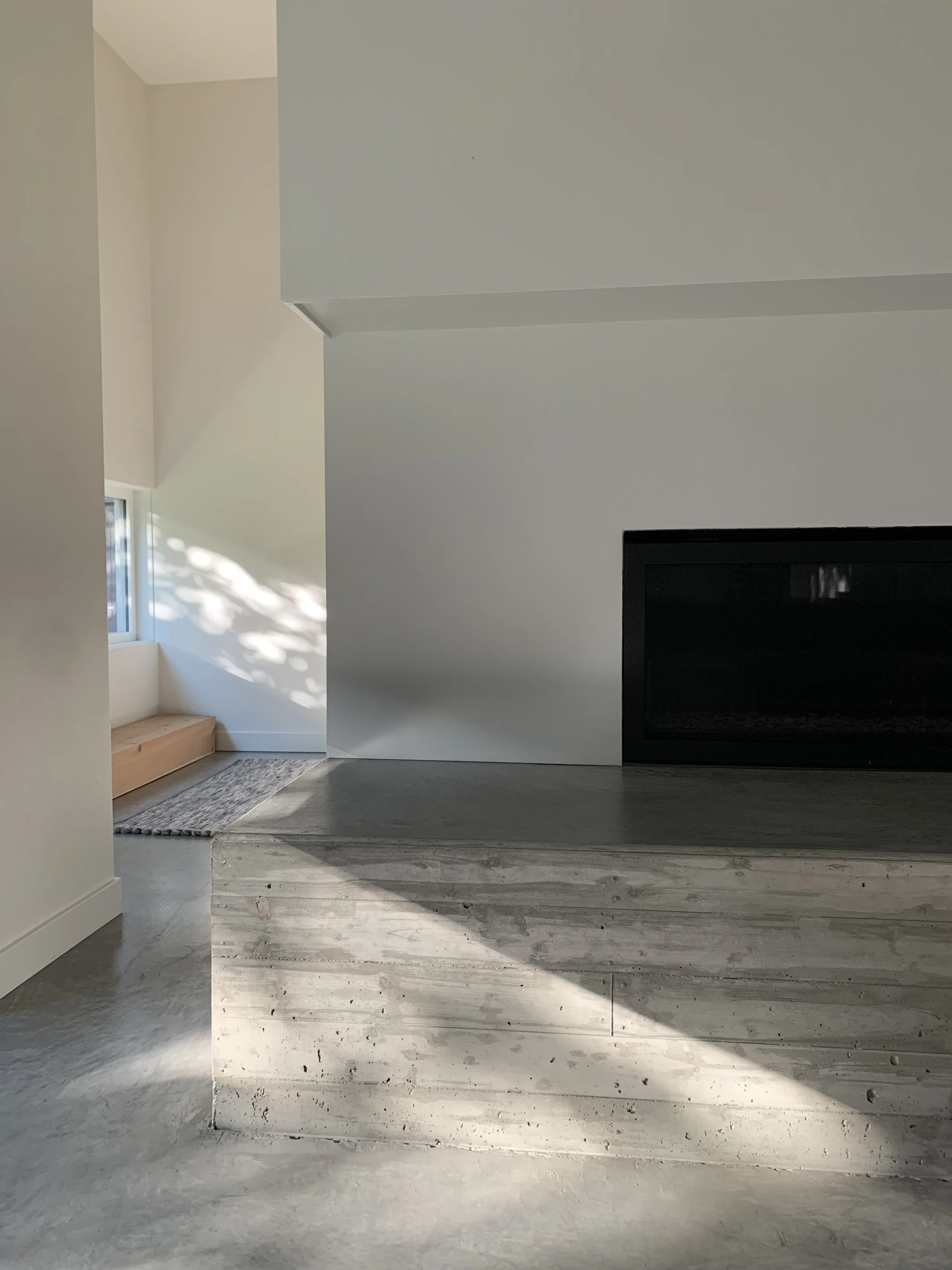 Interior of a modern home with white walls, a small window with sunlight creating shadows, and a black fireplace built into a wooden platform with horizontal wood grain.