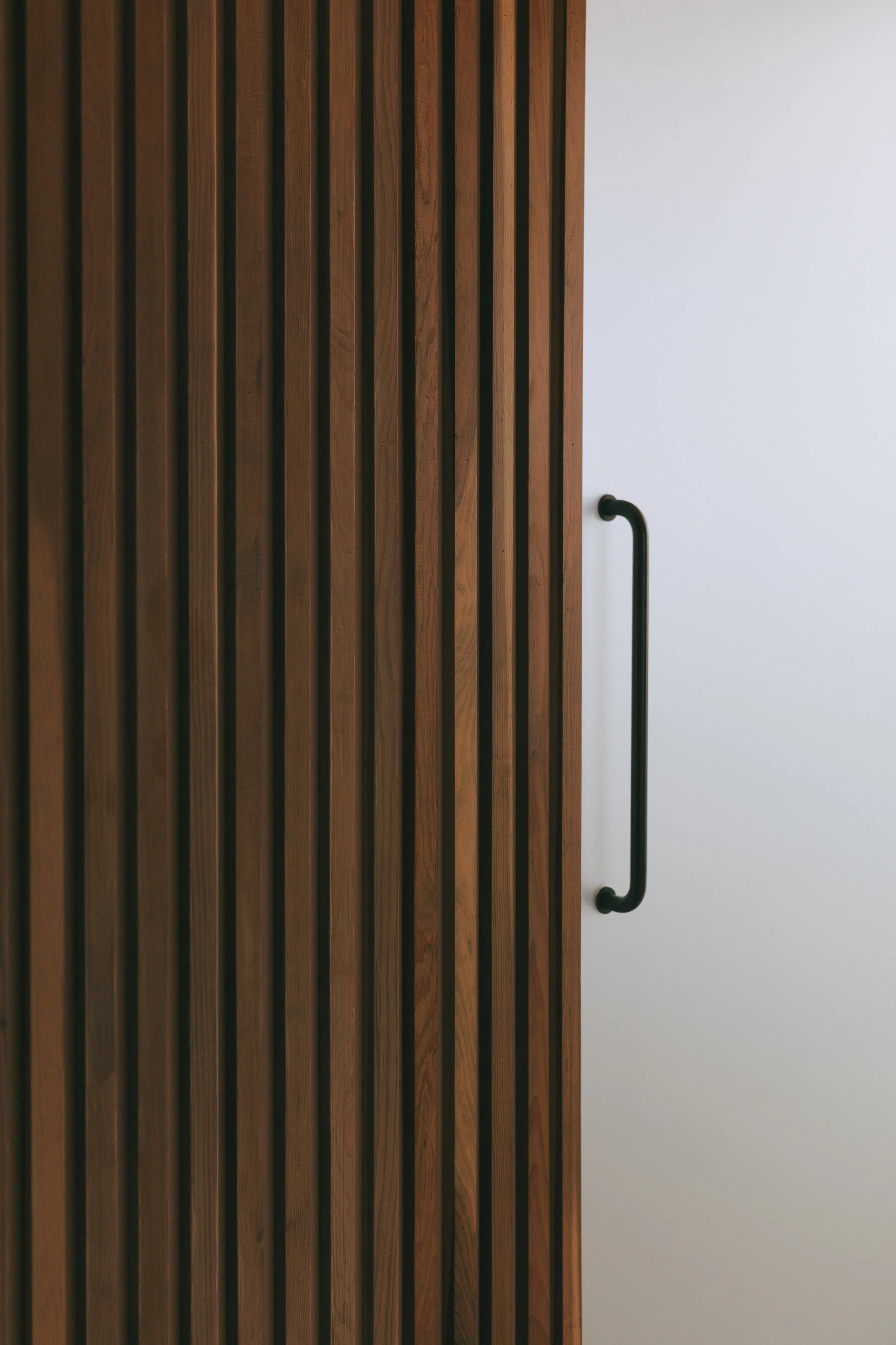 Wood slat door with black handle on the right side of a plain white wall.