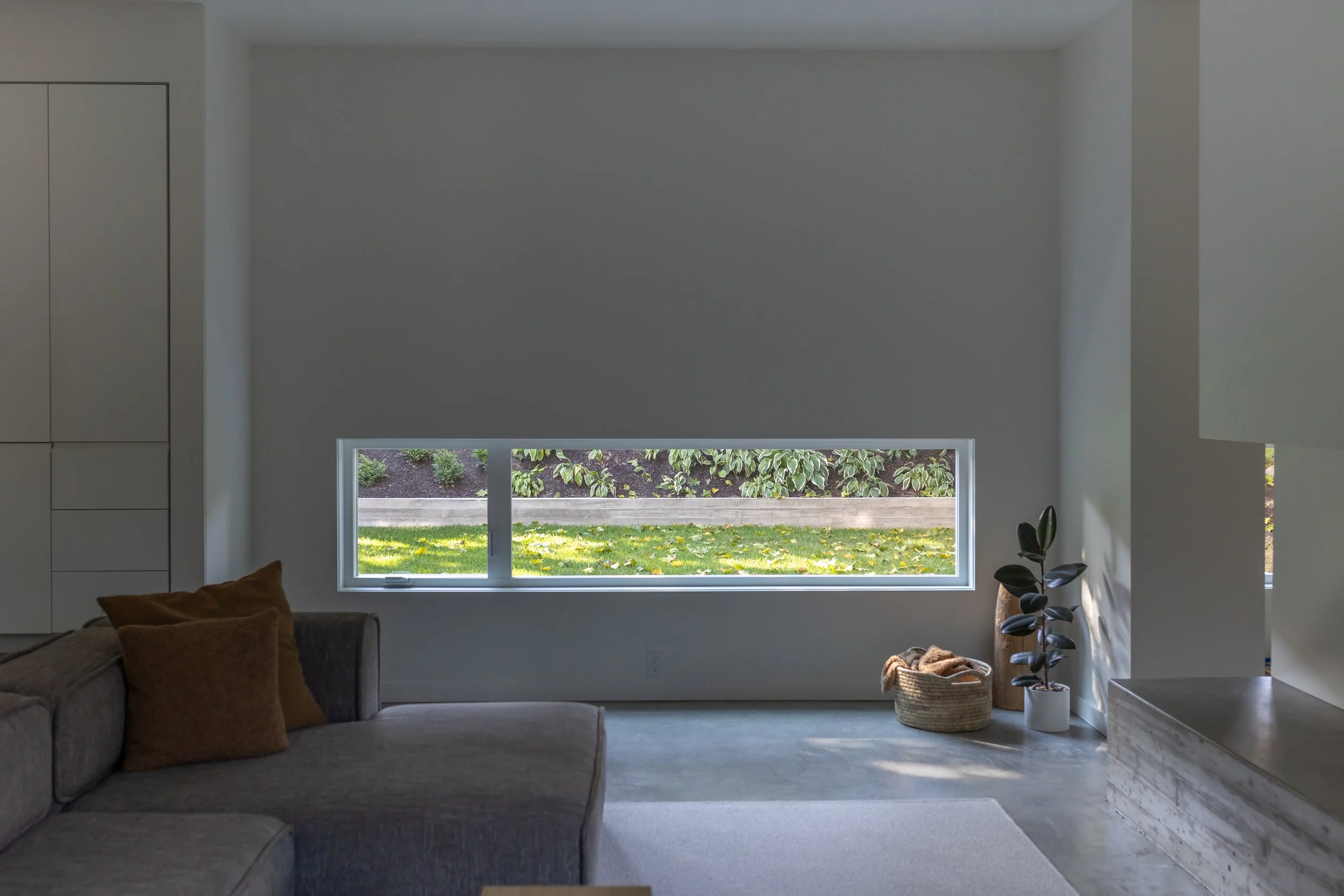 Modern living room with a large window overlooking a garden, gray sectional sofa with brown pillows, indoor plants in baskets and pots, and a marble fireplace.