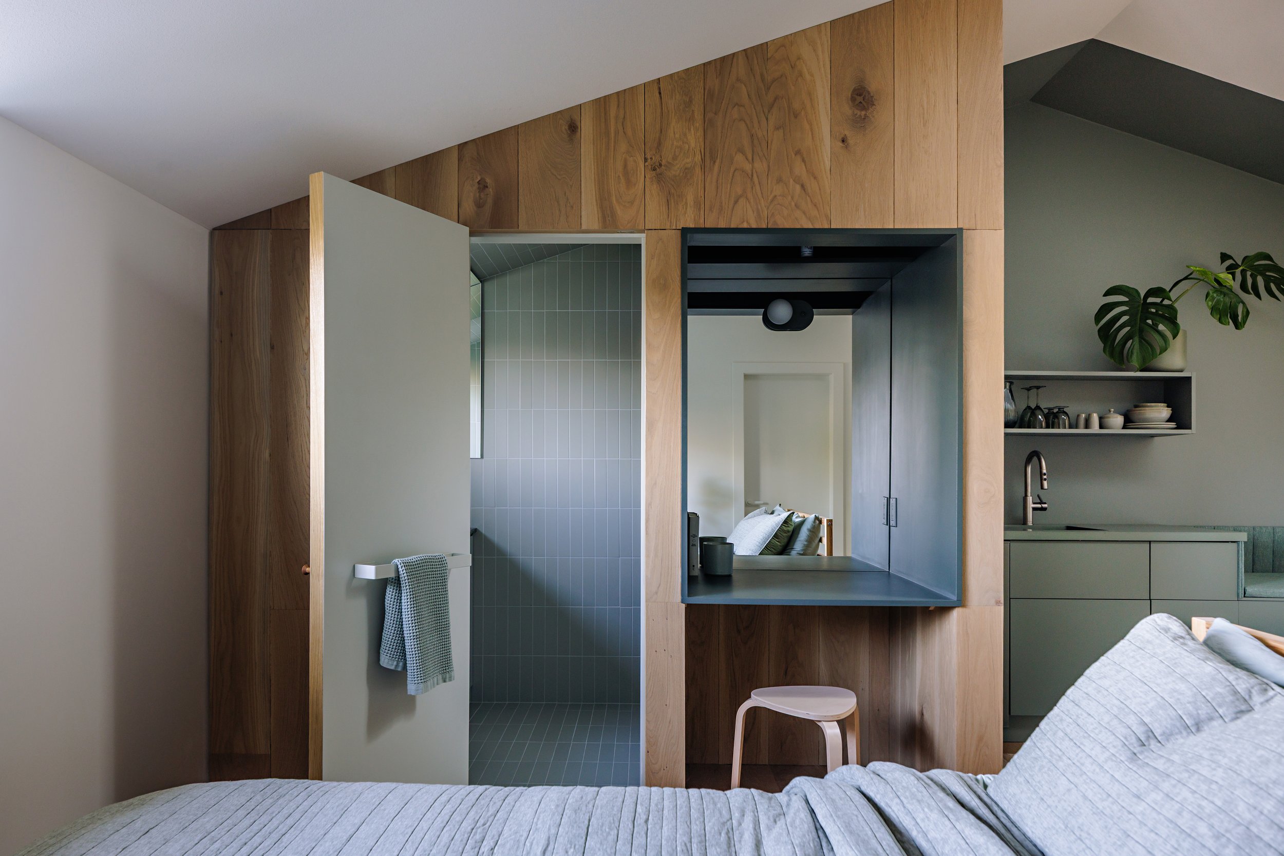 Interior of a modern bedroom with a bed, wooden paneling on the wall, a mirror, a small stool, and a kitchen area with a plant and shelves.