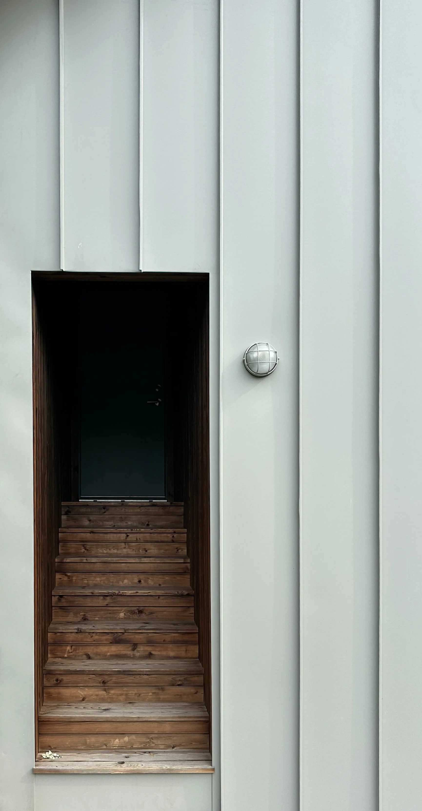 Modern exterior wall with a small recessed entrance featuring wooden steps, a black door, and a metal globe-shaped light fixture.