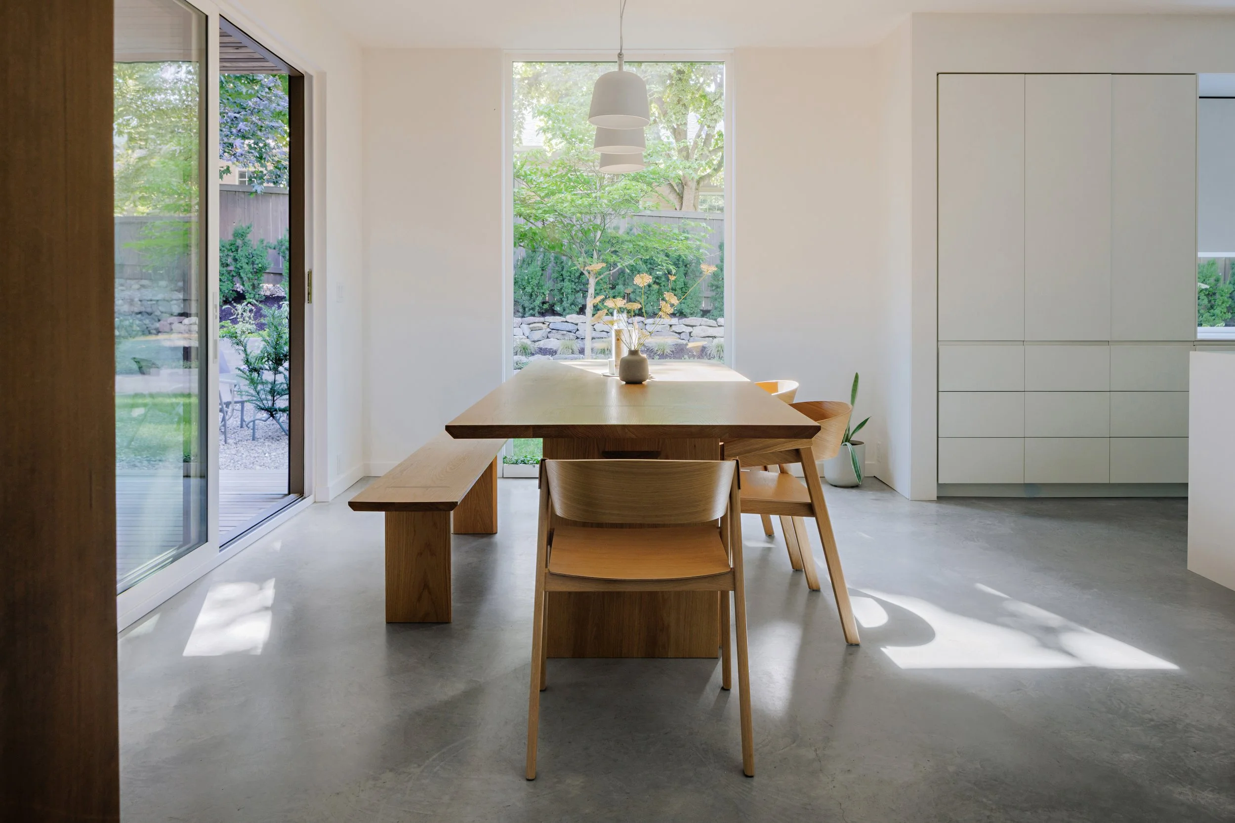 A minimalist dining room with a wooden table and chairs, large windows showing a garden outside, and modern pendant lighting
