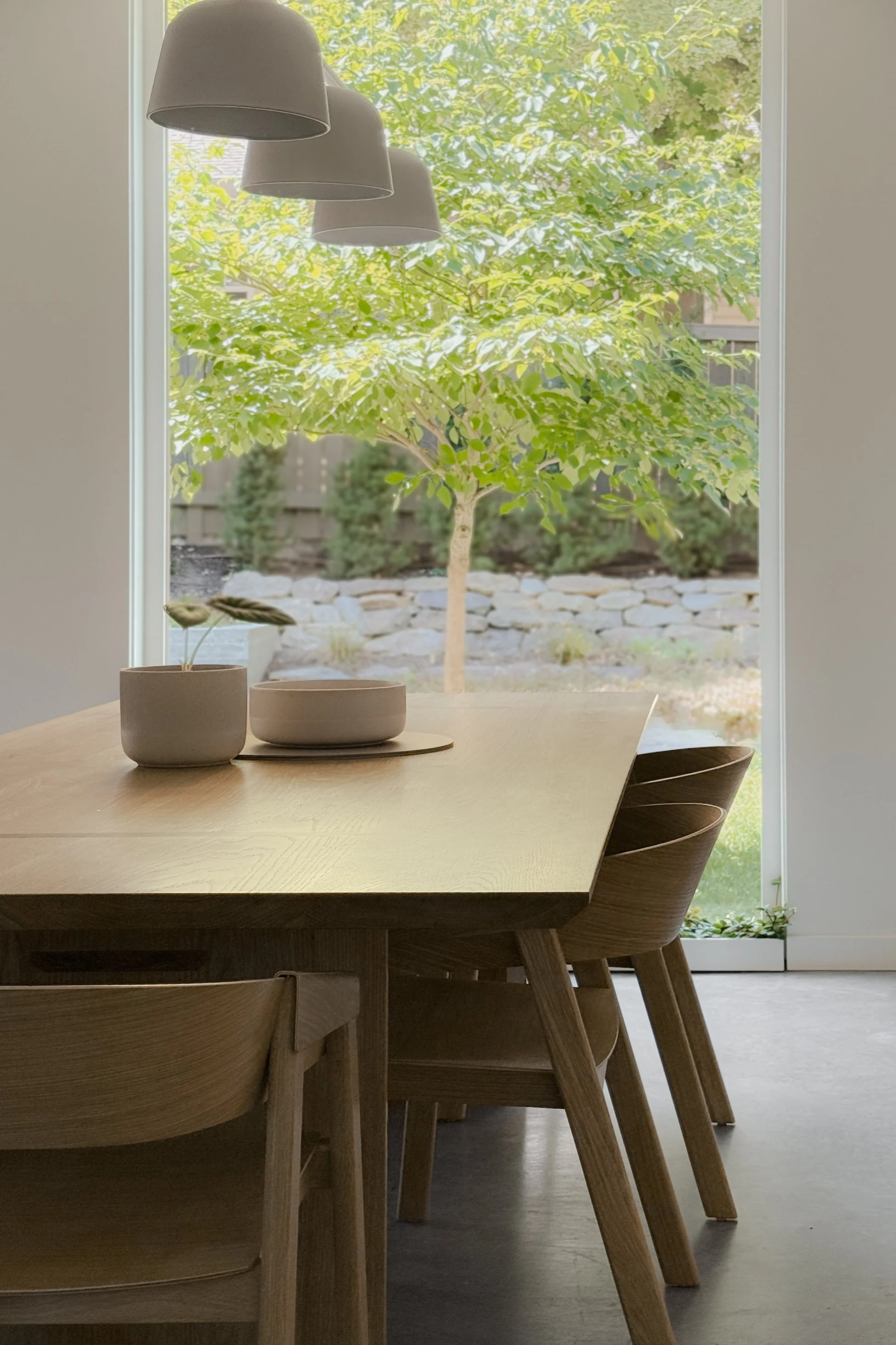 A dining room with a wooden table and chairs, decorative bowls, and potted plants, with a large window overlooking a lush green backyard with trees and stone-edged garden beds. Three ceiling pendant lights hang above the table.