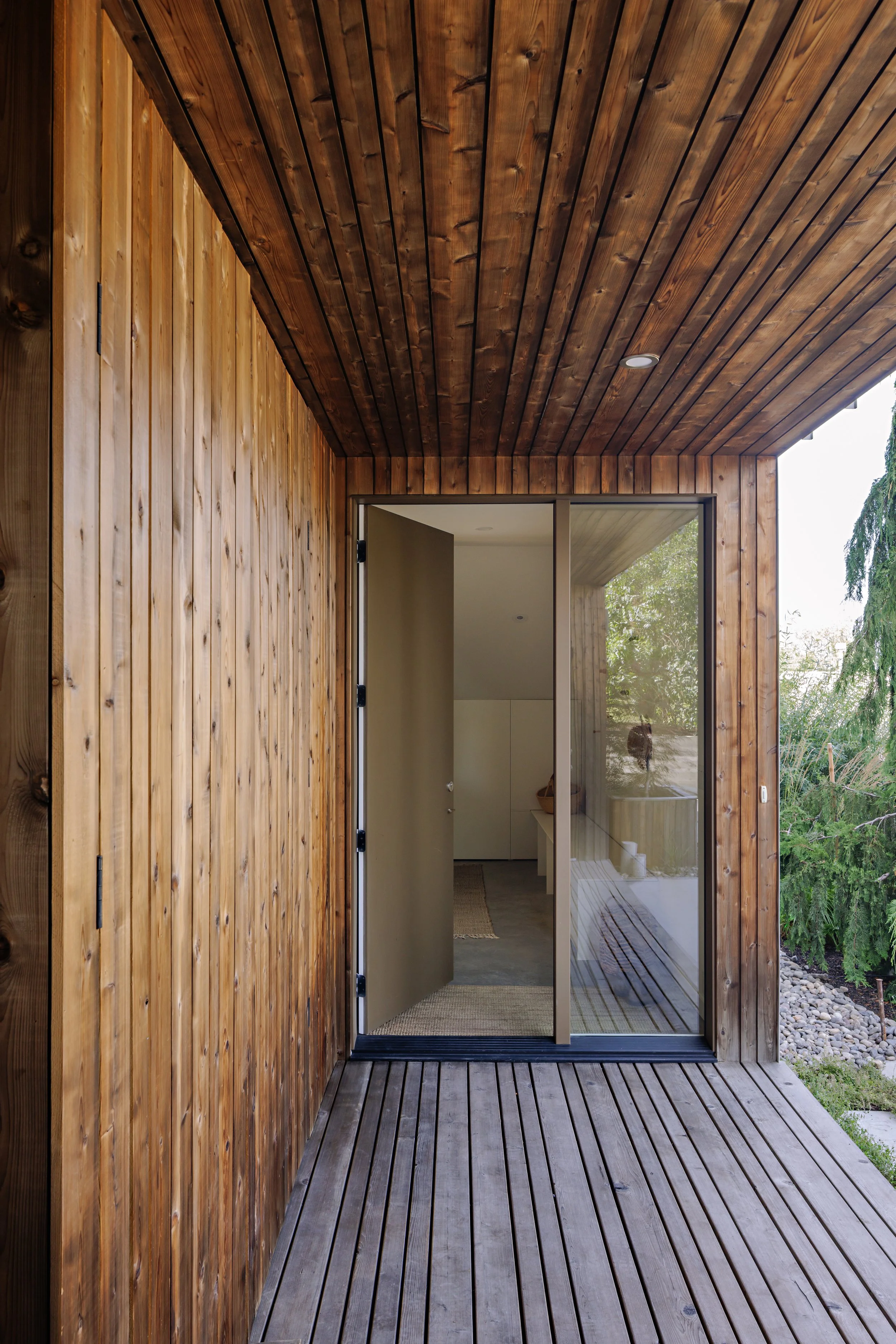 A wooden porch with vertical and horizontal wooden planks, an open door leading to interior space, and outside greenery visible through a large glass window.