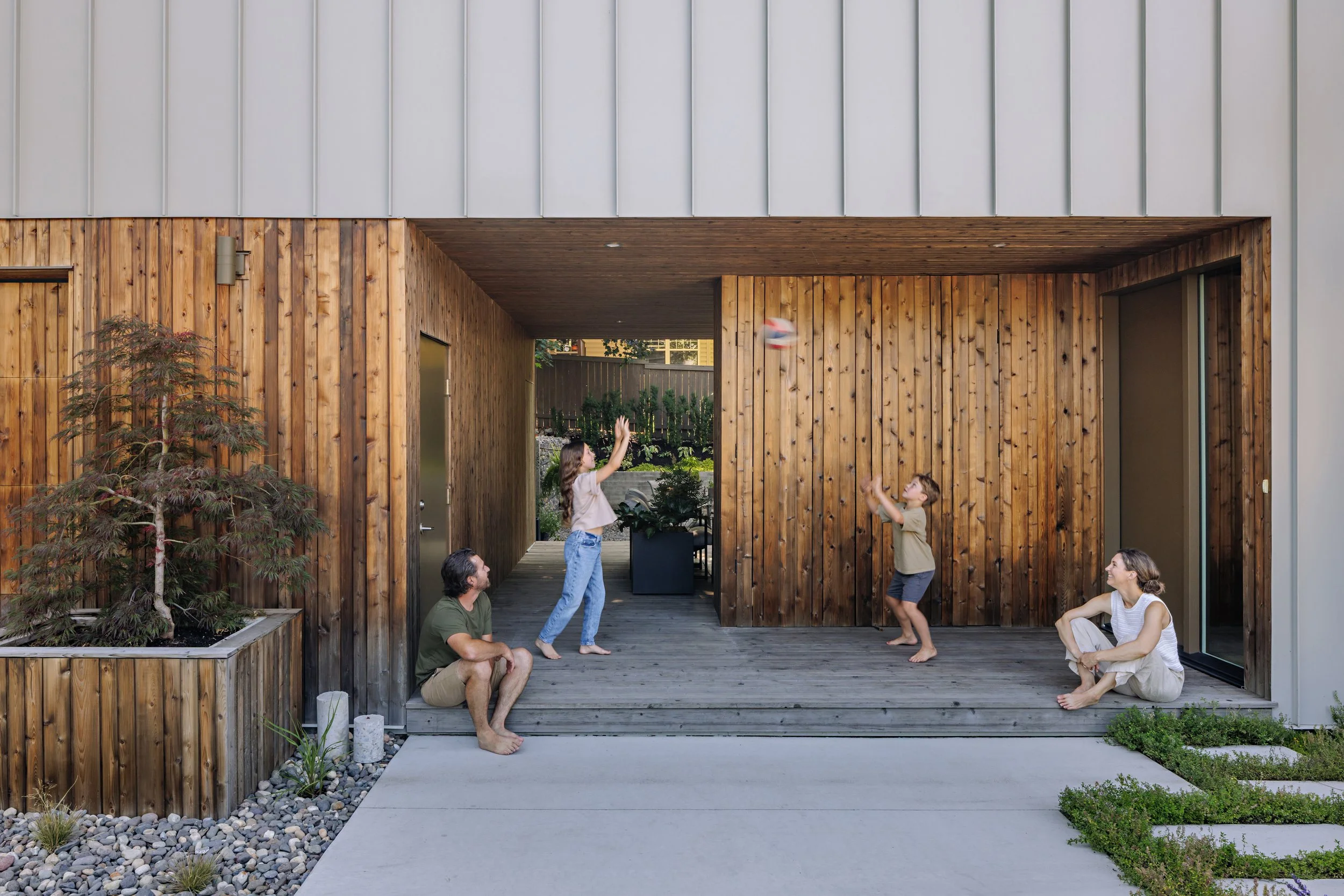 Children and adults playing volleyball on a wooden porch with modern house exterior, featuring wooden and metal panel walls, and nearby plants.