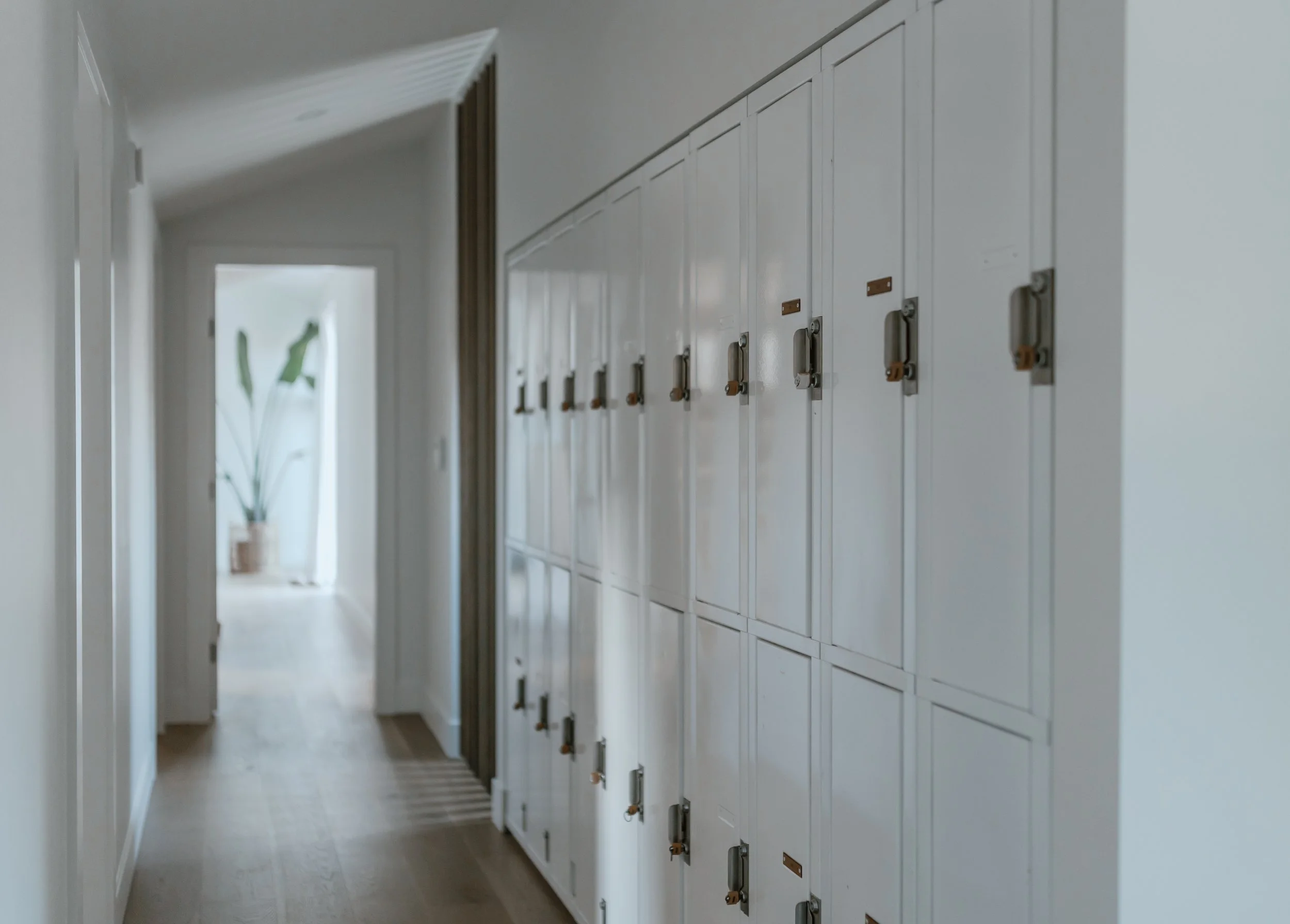 School hallway with white lockers on the right and a doorway at the end, with a potted plant visible in the distance.