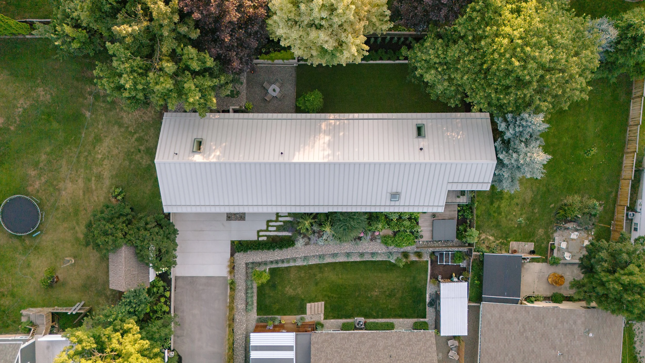 An aerial view of a backyard with a large white metal roof house, a concrete driveway, a trampoline, a gravel path, a garden area with plants, and surrounding greenery including trees and lawn.
