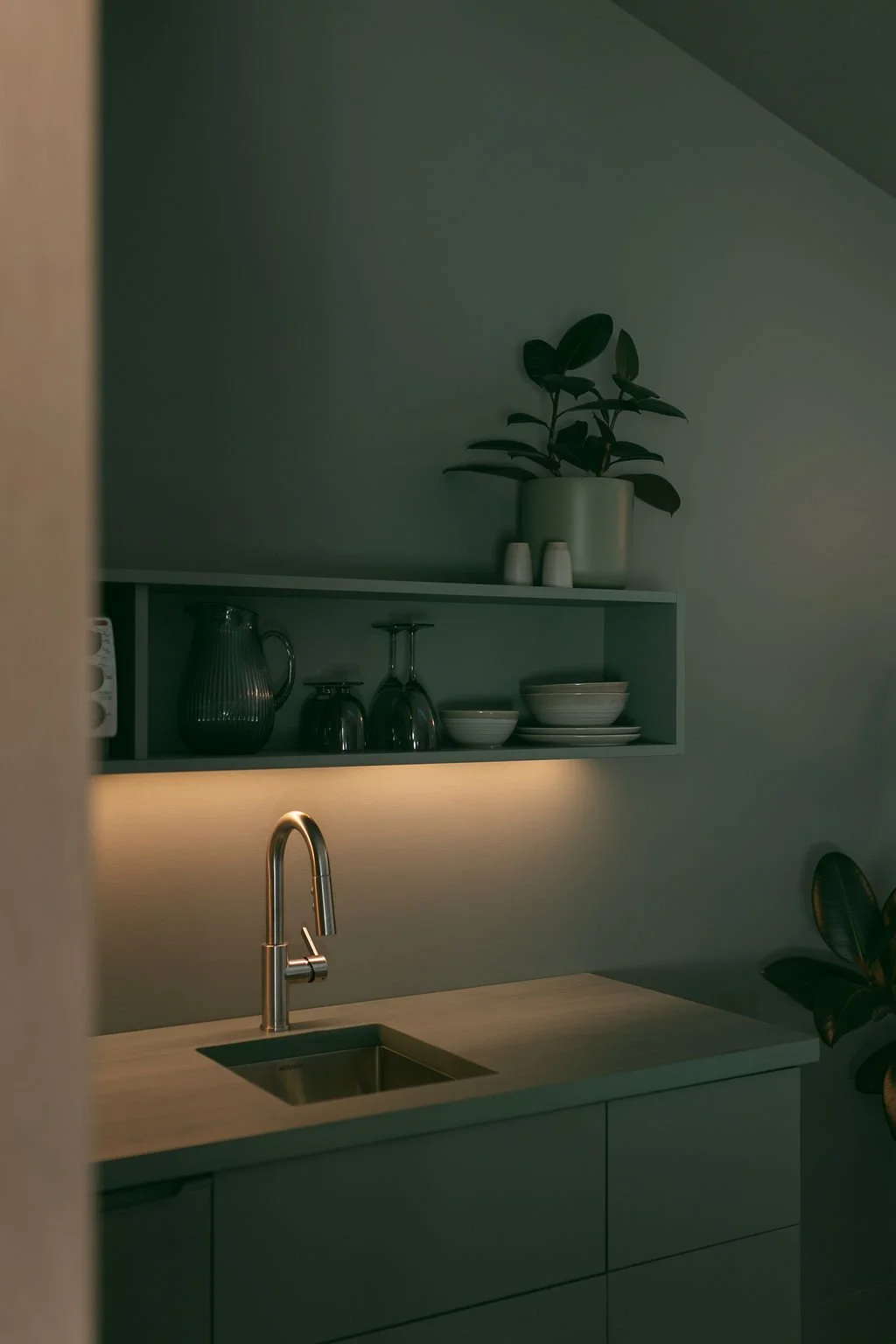 A minimalist kitchen with a light wooden countertop, a stainless steel faucet, a sink, a green open shelf with bowls, glasses, and pitchers, and a potted plant on the wall behind.