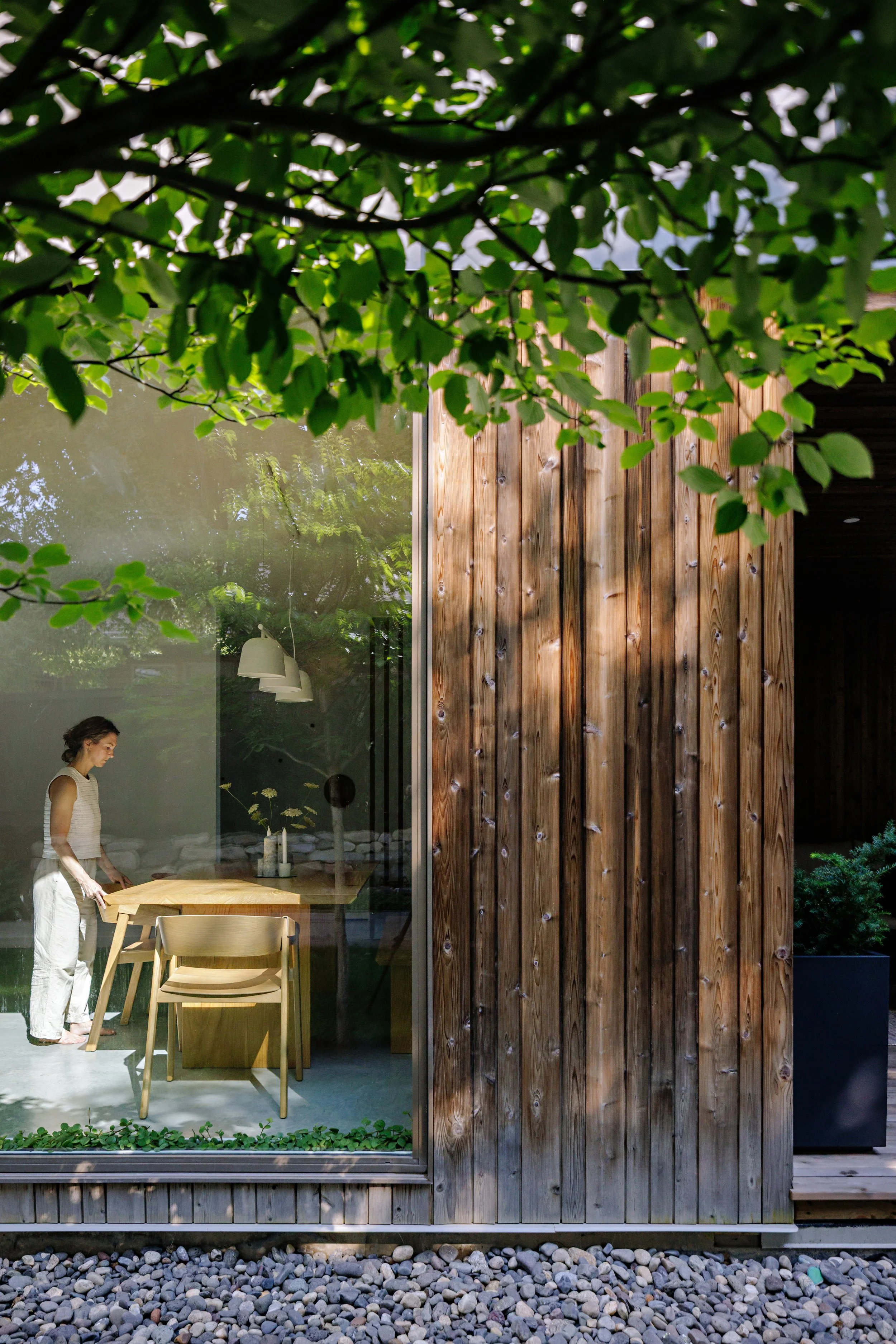 A woman with dark hair tied back, wearing a sleeveless top and white pants, standing inside a home next to a wooden dining table with a modern design. The interior is visible through a large glass window, with a wooden wall outside, a window reflecti