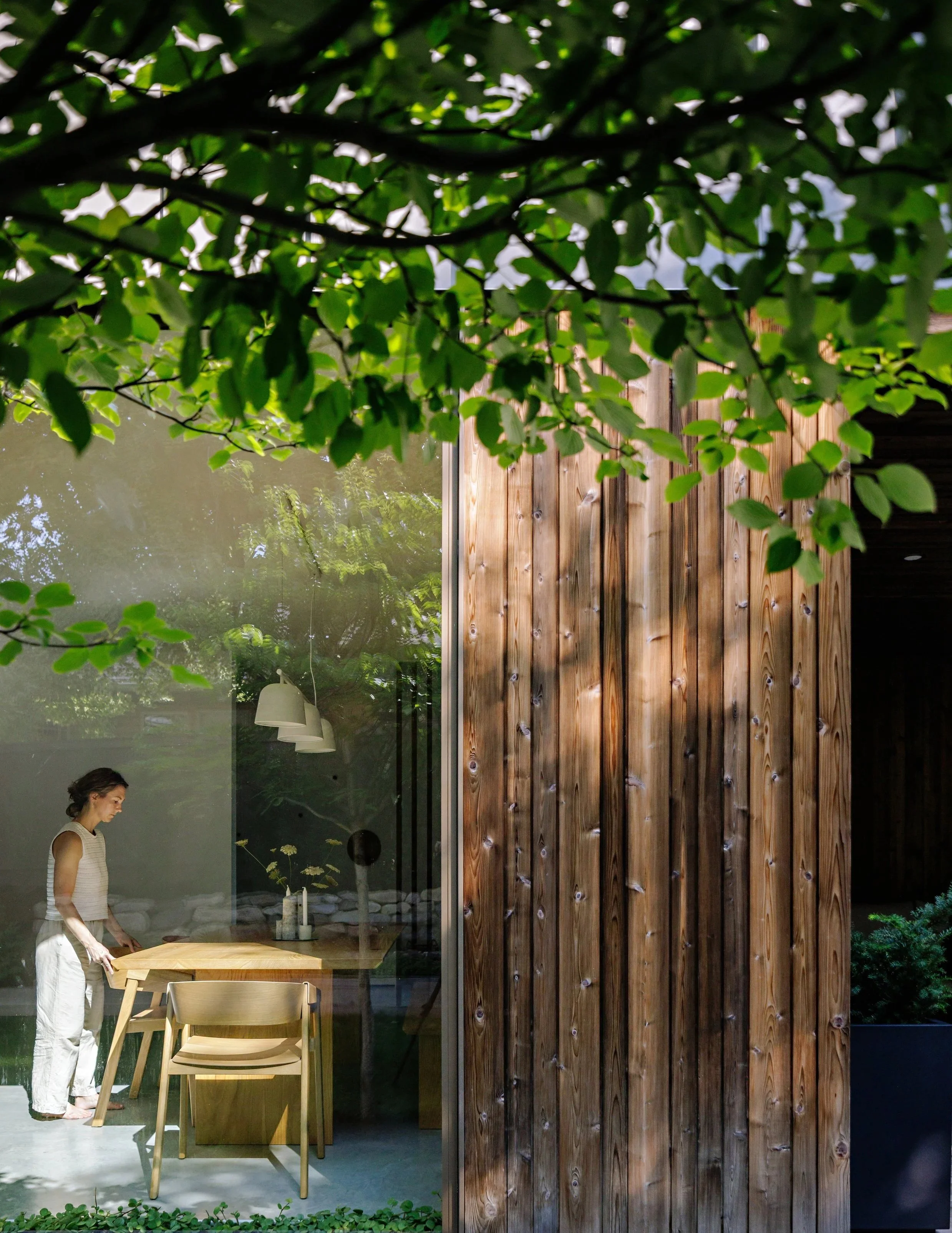 A woman in a sleeveless top and white pants is standing inside a house near a wooden dining table with chairs, visible through a large glass window with a reflection of trees and sky outside.