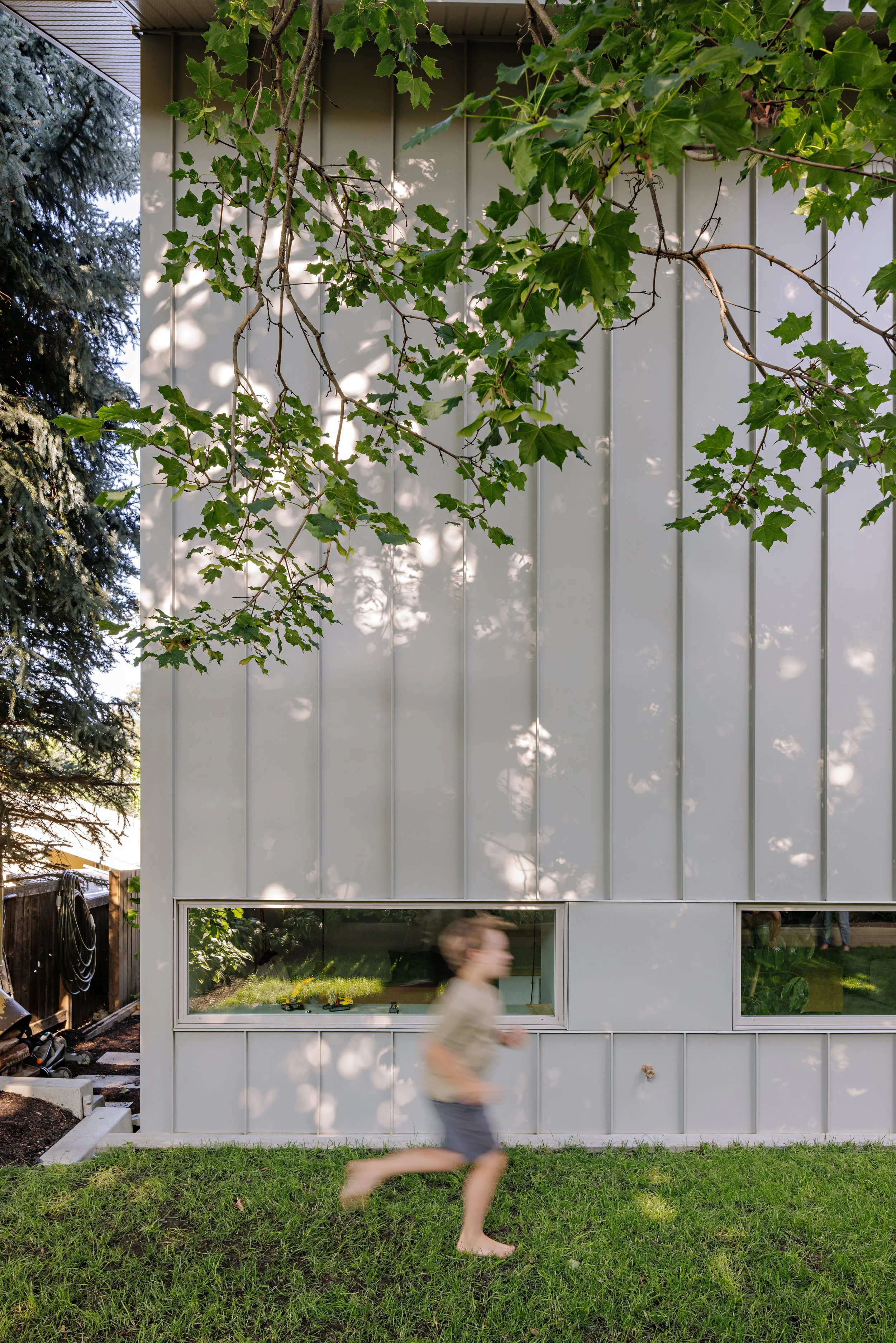 A woman jogging on a lawn in front of a modern house with large windows and white siding, partially shaded by tree branches.