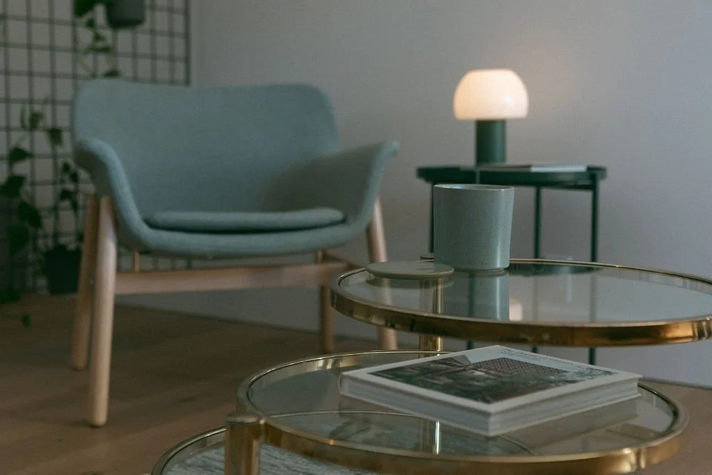 Living room with a light blue armchair, a gold and glass coffee table with magazines, a side table with a lamp, and a decorative wall grid with plants.