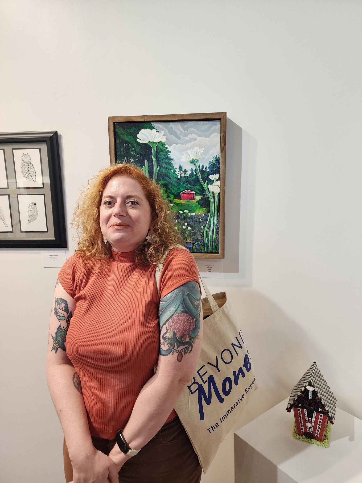 A woman with curly red hair and tattoos on her arms standing at an art exhibit. She wears an orange ribbed shirt, has a black watch on her left wrist, and carries a beige tote bag. Behind her, a colorful painting of white flowers with a red barn in a green landscape is displayed on the wall. To her right, there's a small LEGO model of a red barn on a white pedestal.