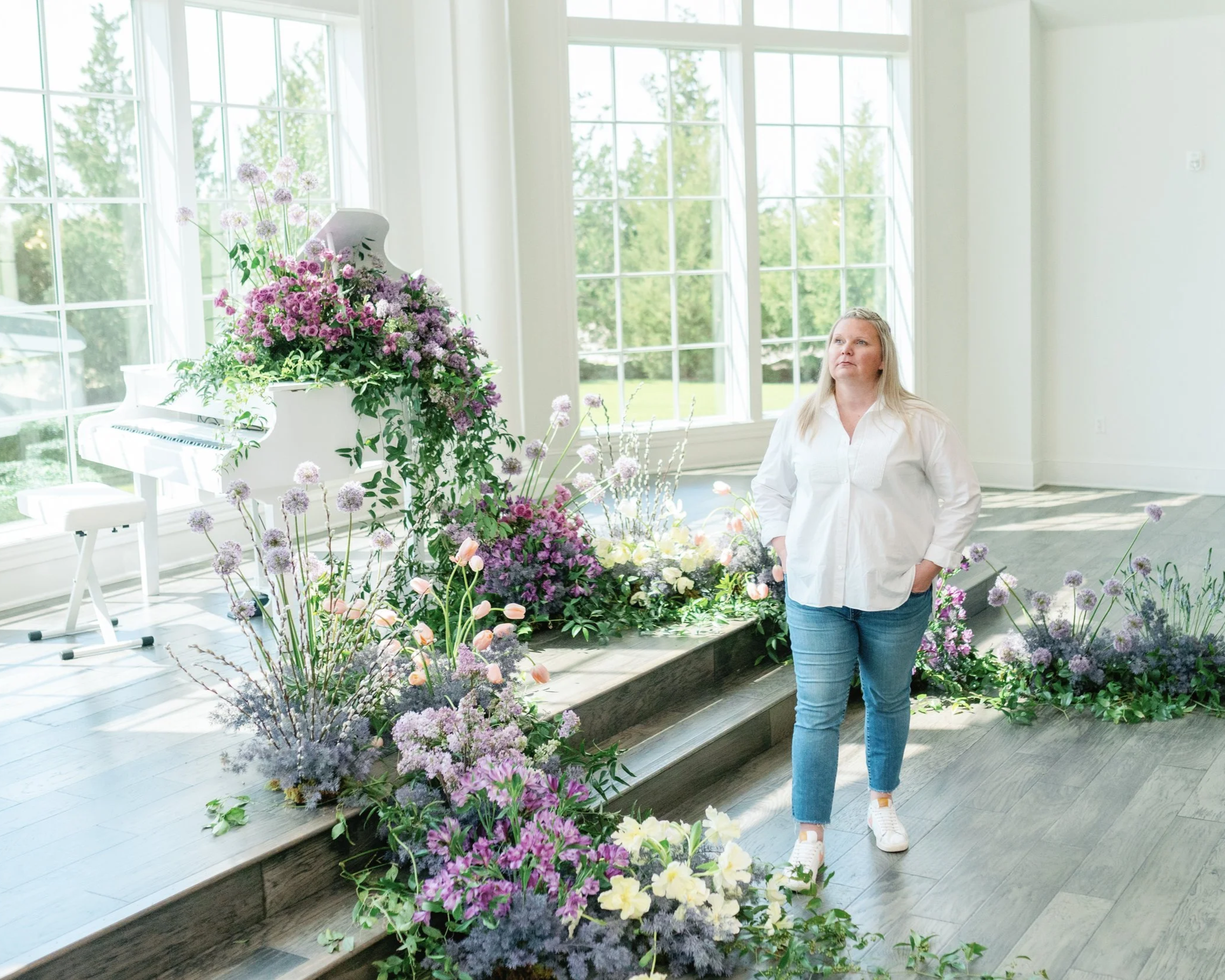 Fashionable Florist standing in front of a wall of windows and a piano surrounded by flowers.