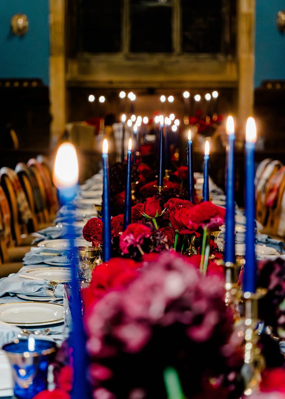 Long rectangular table designed by Fashionable Florist. The table has red and burgundy flowers and a sea of blue taper candles.