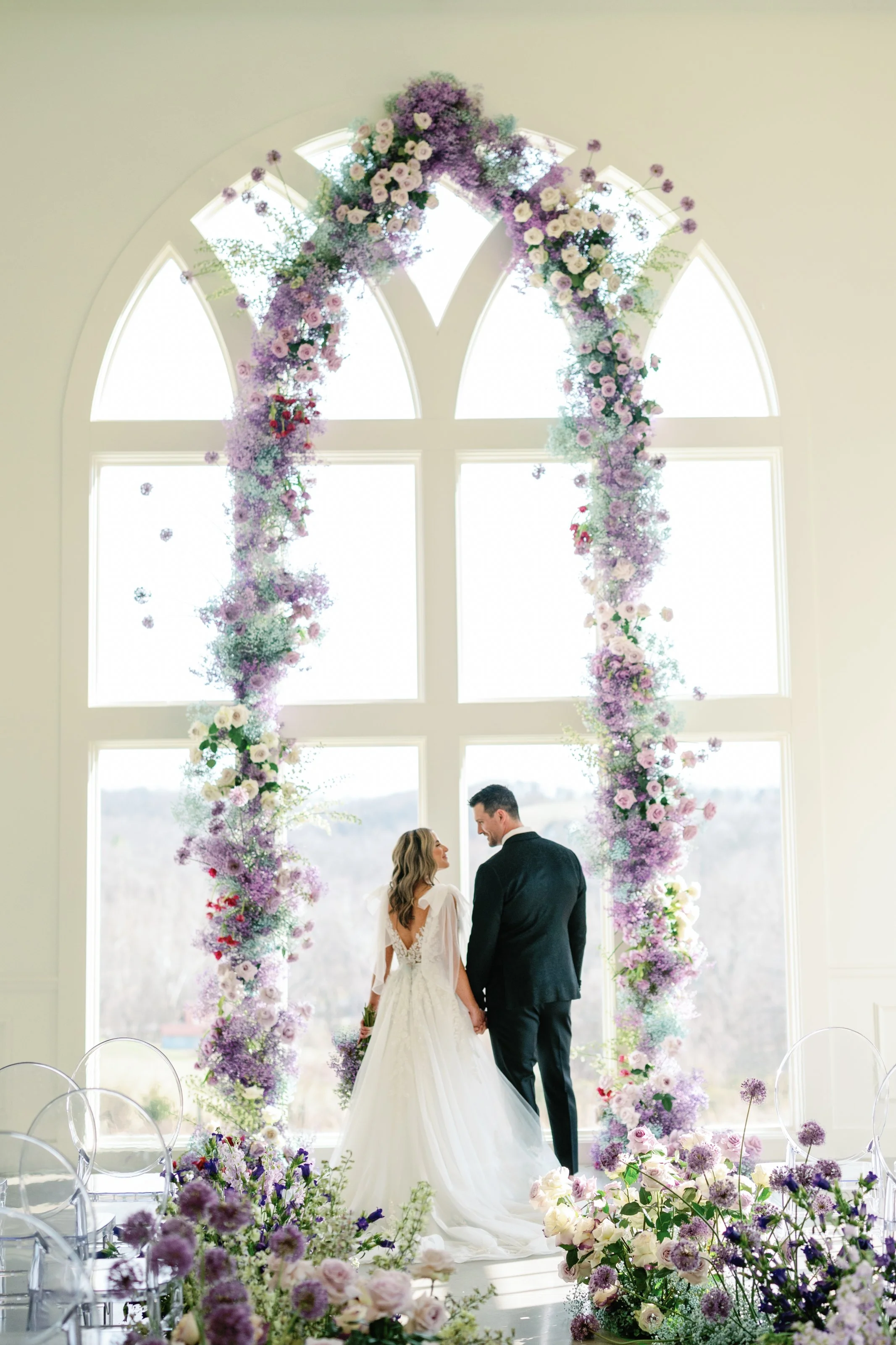 A couple standing in front of a large window with flowers all around them designed by Fashionable Florist.