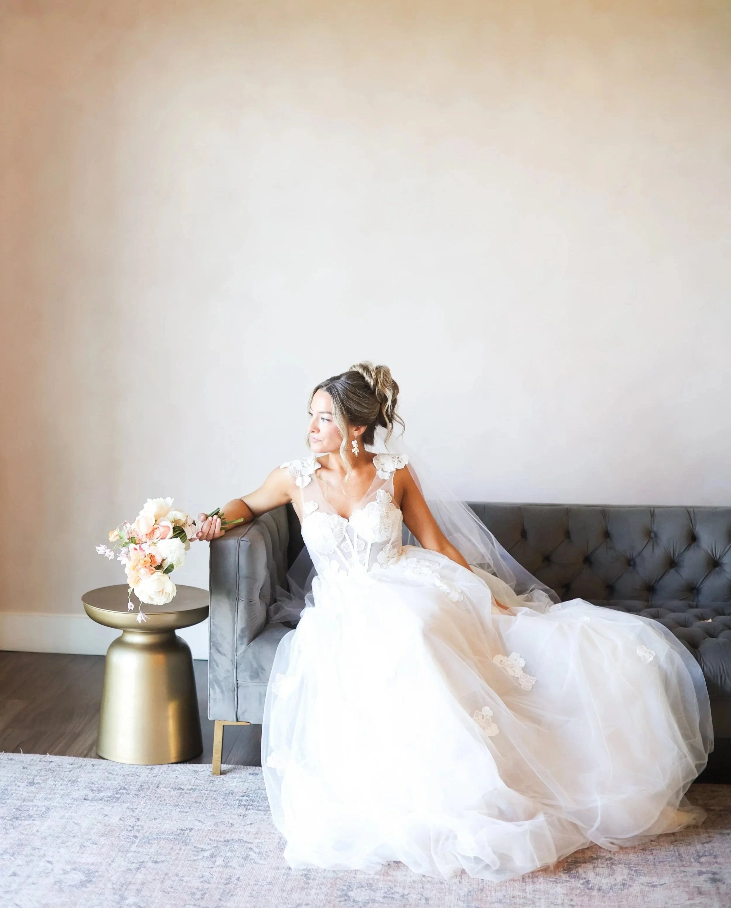 Bride sitting on a gray couch holding a bridal bouquet.