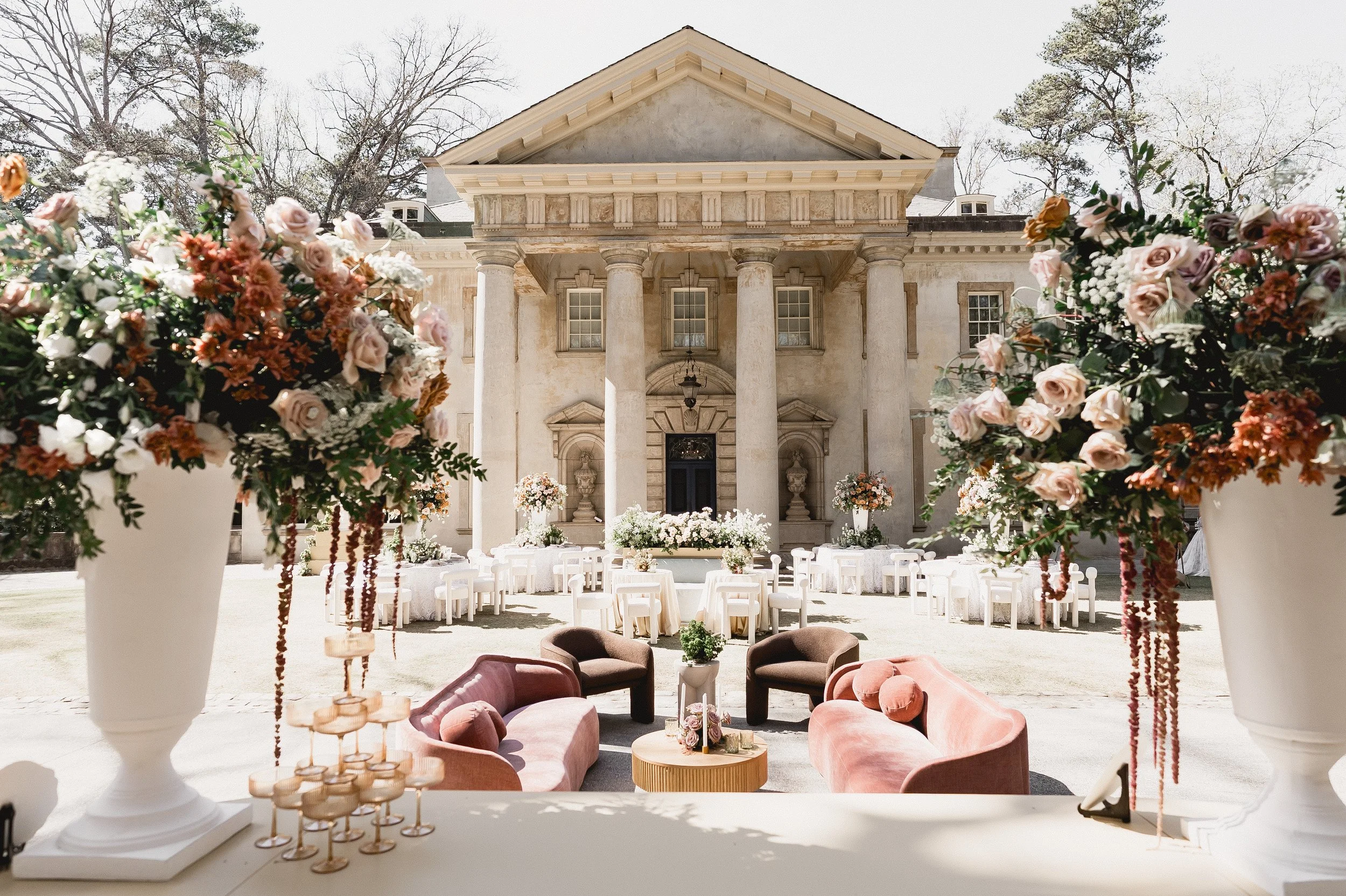 Elegant outdoor wedding setup with soft pink and white floral arrangements, comfortable pink and mauve seating, and a historic building with large columns.
