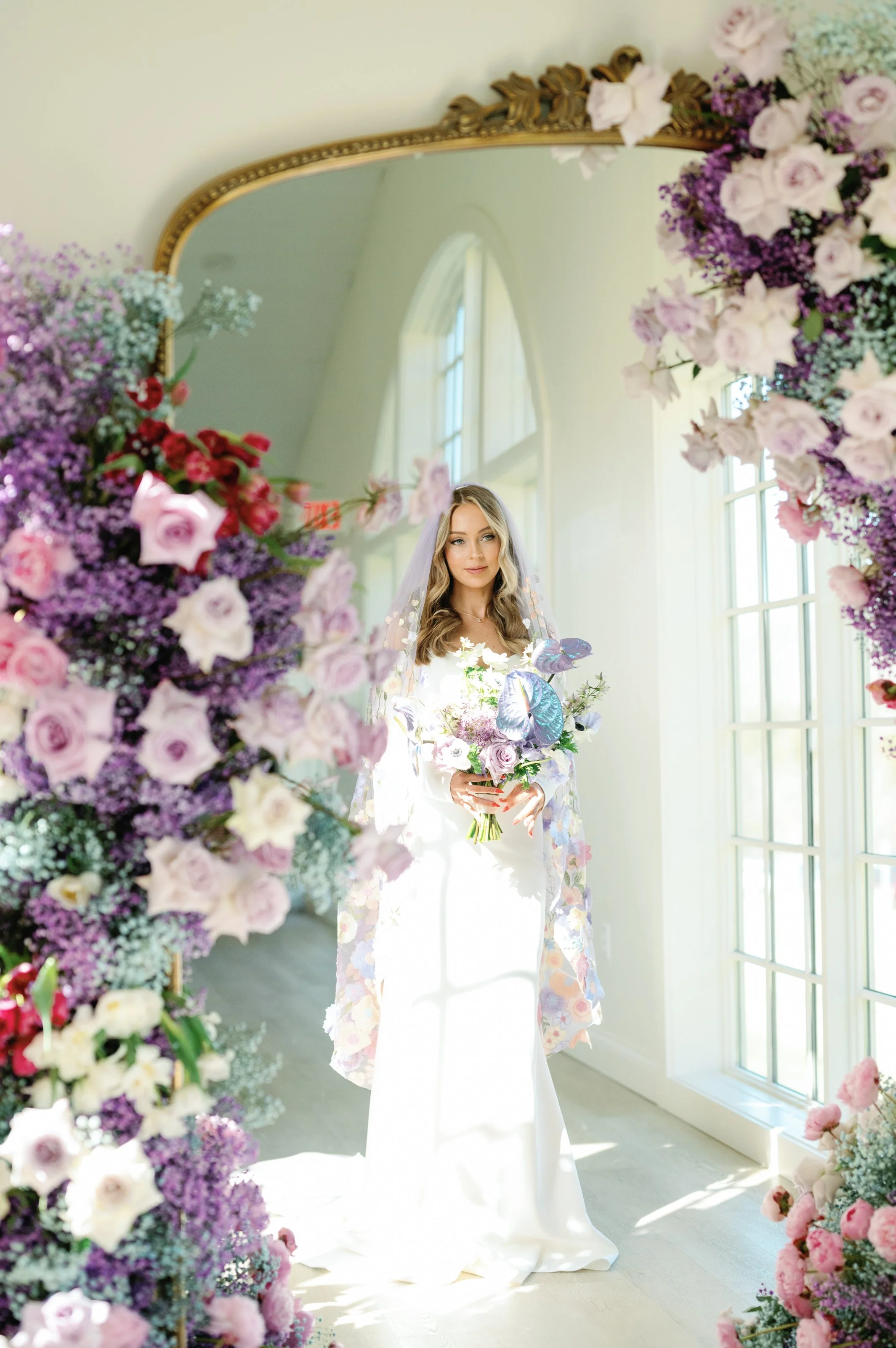 A bride standing in front of a mirror adorned with flowers.