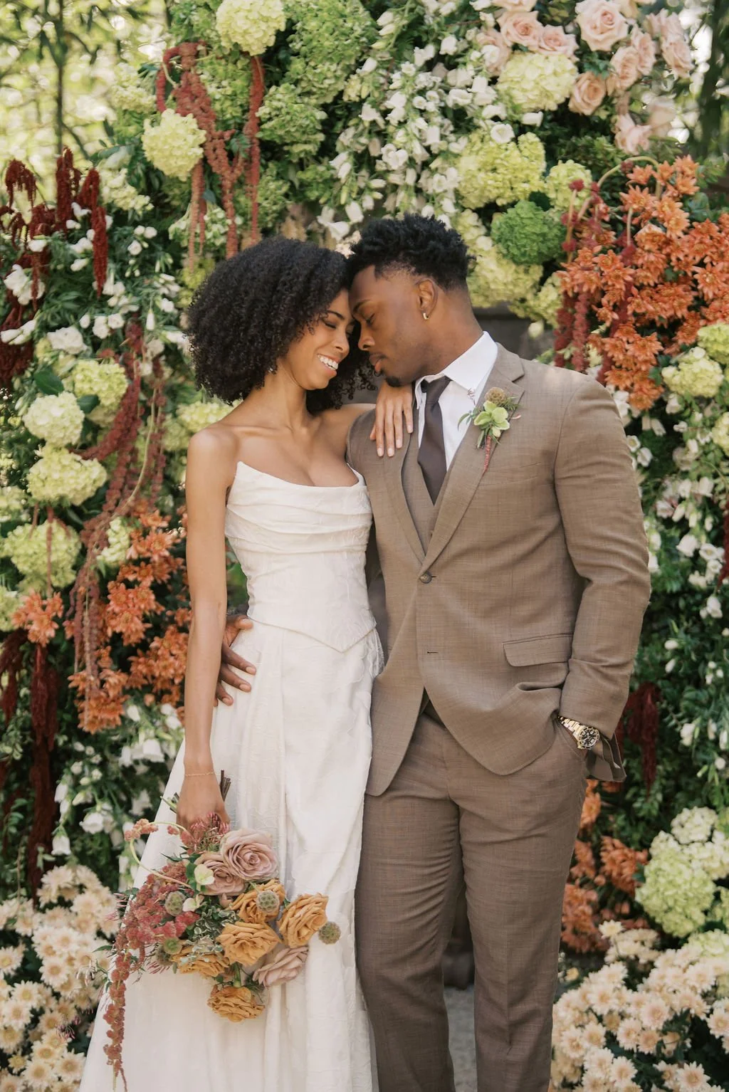 A couple in wedding attire, standing close and touching foreheads, smiling in front of a lush floral backdrop.