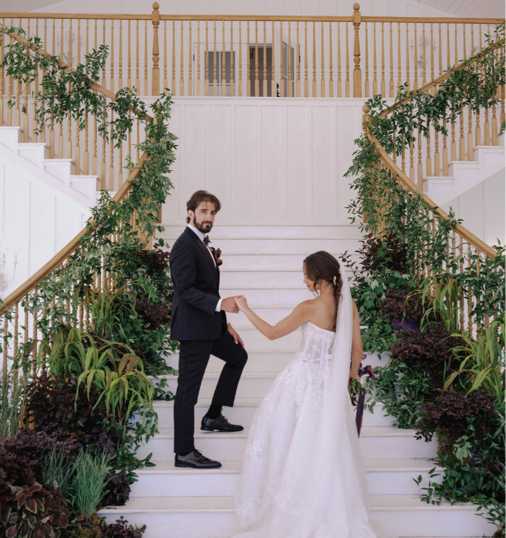 Bride and groom going up a large white staircase that is adorned with green and burgundy plants.
