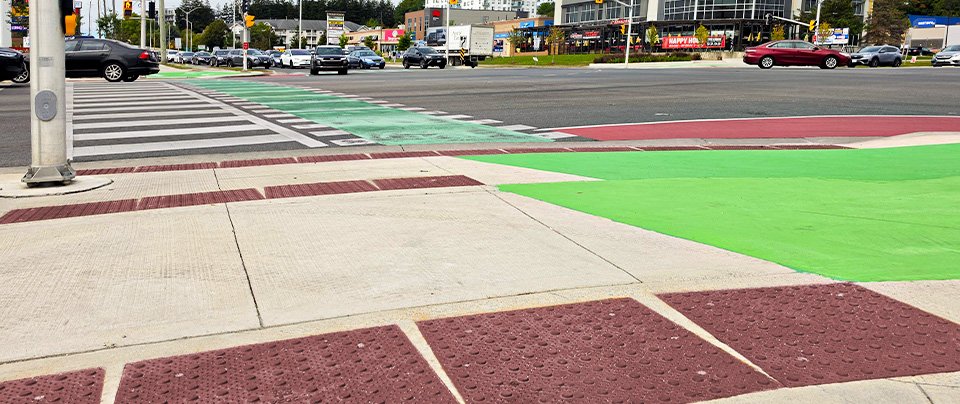 Pedestrian crosswalk at a traffic intersection with GORA brick red steel tactile and radial plates leading up to the crossing with both green bicycle lanes and red bus transit lanes.