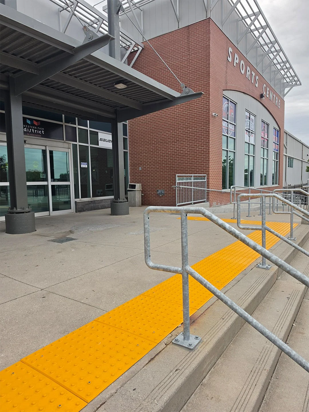 Exterior view of the Western Fair Sports Centre in London Ontario, a concrete stairway leads up to the building with GORA removable 24x24" safety yellow fiberglass tactile plates sitting at the top of the stairway.