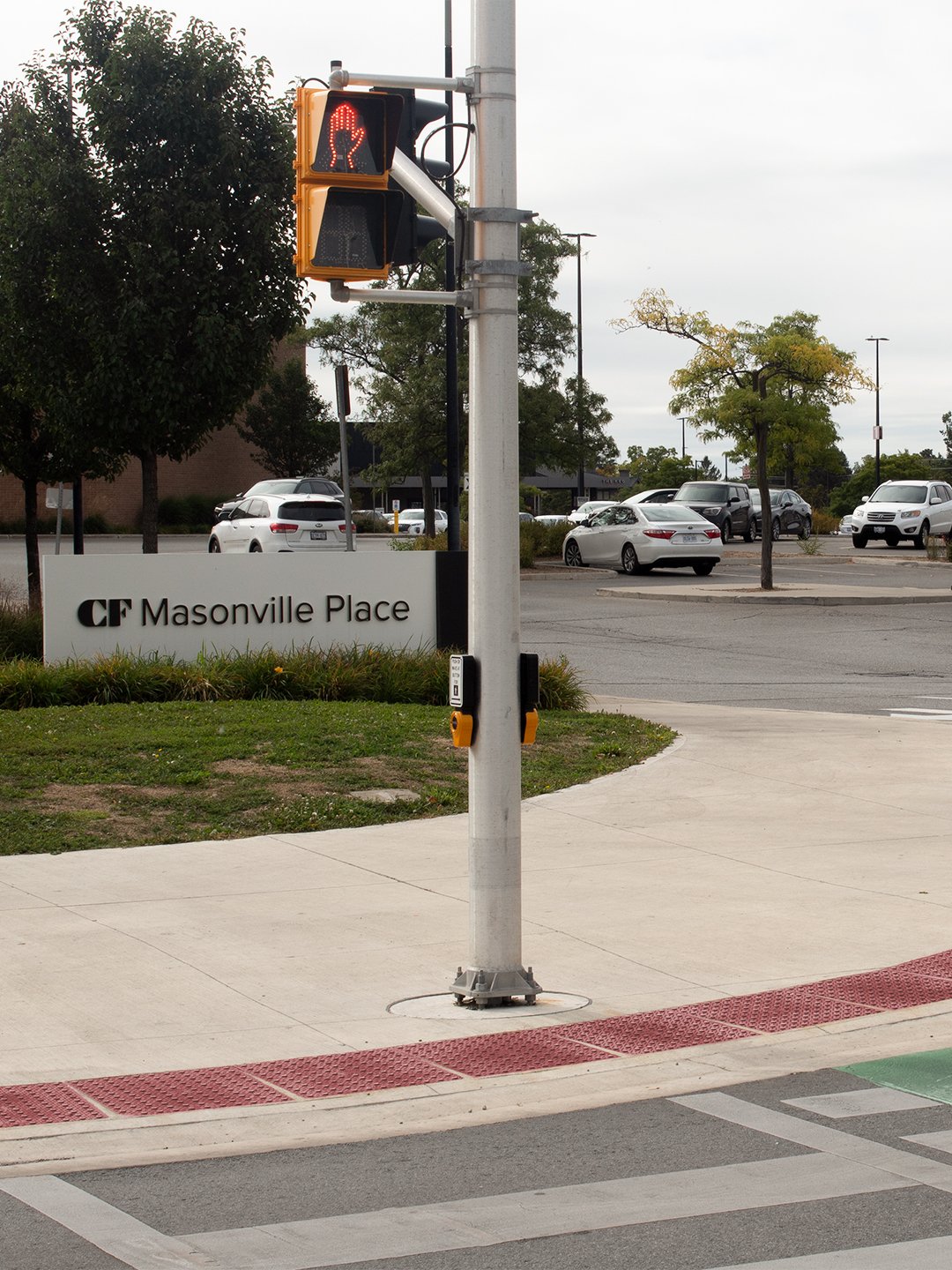 Pedestrian crosswalk leading up to Masonville Place Shopping Mall, the sidewalk has GORA brick red steel tactile and radial plates embedded into the concrete just before the intersection.