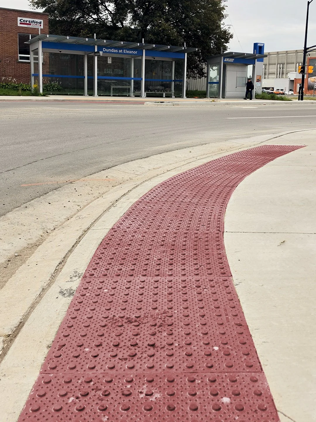 A pedestrian crosswalk at Dundas and Burbrook in London Ontario, with GORA brick red steel tactile and radial plates on the sidewalk just before the intersection begins.