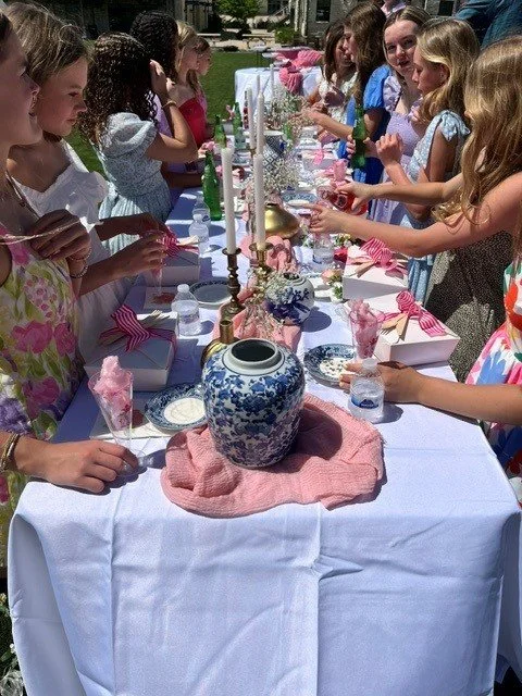 Children gathered around a long table set for a celebration, with pink and white decorations, candles, and a blue and white porcelain vase in the center.