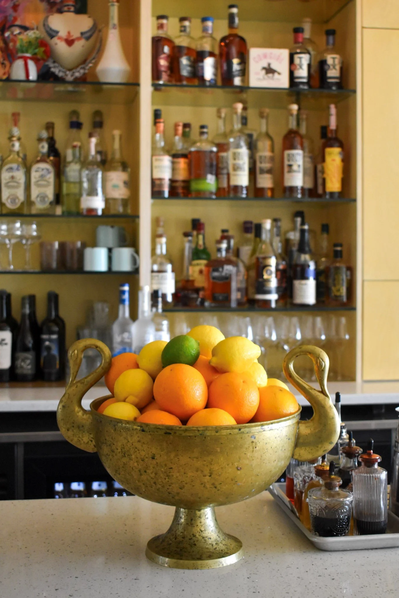 A decorative gold-colored bowl filled with oranges, lemons, and lime on a kitchen countertop. In the background, there is a glass-fronted cabinet with bottles of alcohol and various kitchen items.
