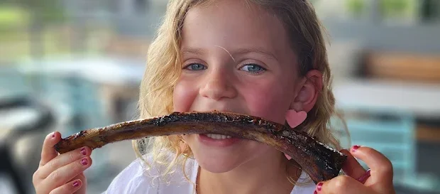 A young girl with blonde curly hair and pink heart-shaped earrings holding a large piece of grilled or barbequed meat in her mouth outdoors.