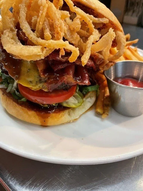 Close-up of a burger with onion rings, lettuce, tomato, bacon, and sauce, served with a side of French fries and ketchup
