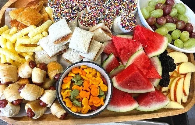 Assorted fruits and snacks on a wooden platter, including watermelon slices, grapes, apple slices, marshmallow pops with sprinkles, fruit-flavored marshmallow candies, and a bowl of mixed gummy candies.
