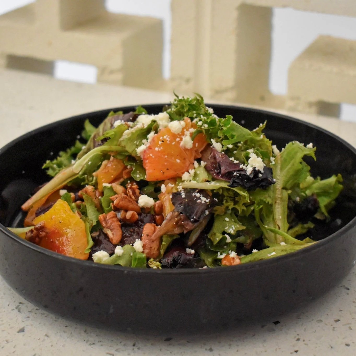 Mixed green salad with chunks of pumpkin, nuts, and feta cheese in a black bowl on a white surface.