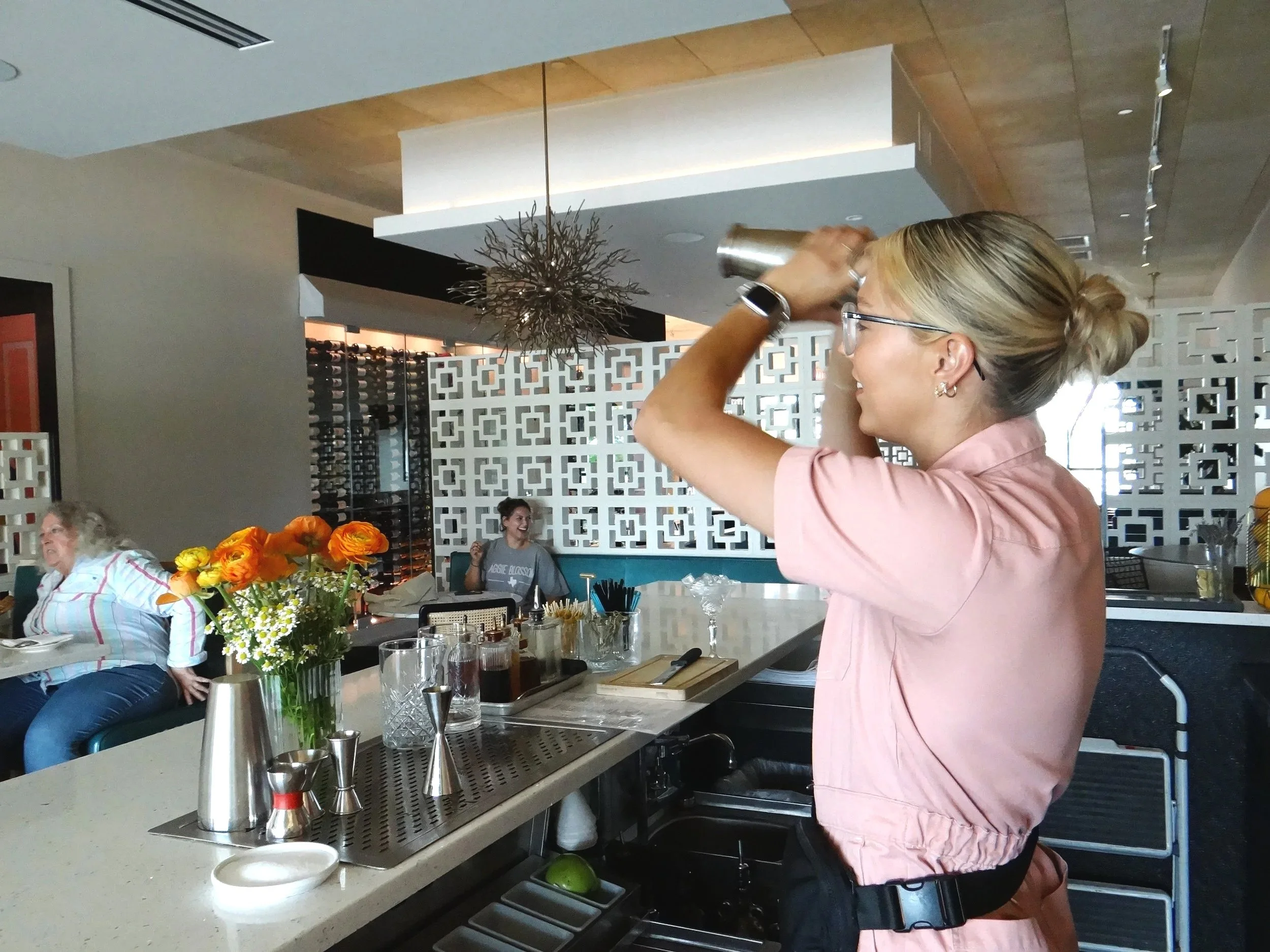 A woman with blonde hair tied in a bun, wearing glasses and a pink shirt, is pouring a drink from a metal shaker. The setting is a restaurant or bar with a counter, glasses, and a flower arrangement on the counter. There are decorative dividers and wine bottles in the background.