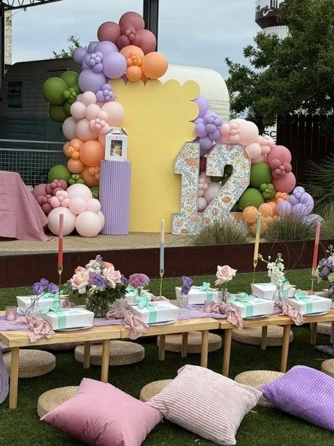 Decorative outdoor setup for a 12th birthday celebration featuring pink, purple, peach, and green balloon arrangements, a large number 12, and a table with wrapped gifts surrounded by cushions.