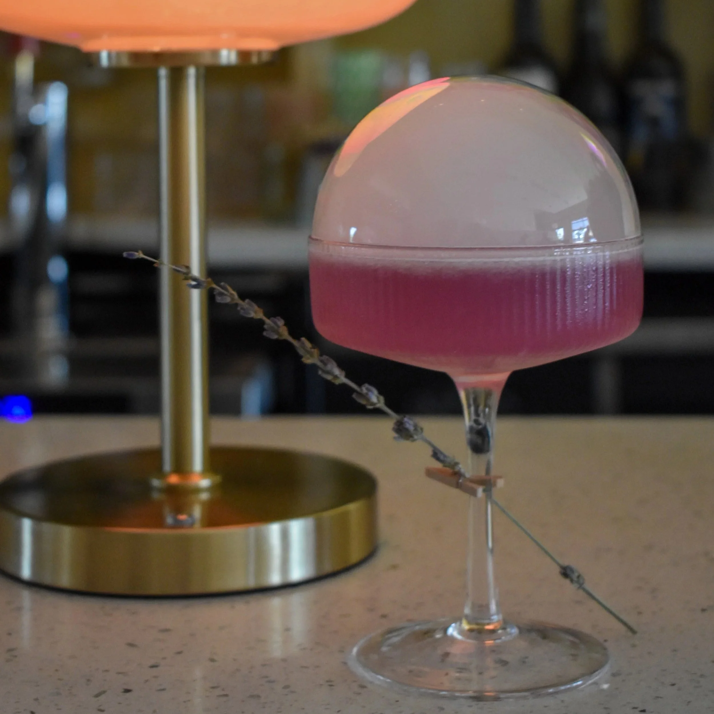 Close-up of a pink cocktail in a stemmed glass with a dome-shaped lid, sitting on a speckled beige countertop.