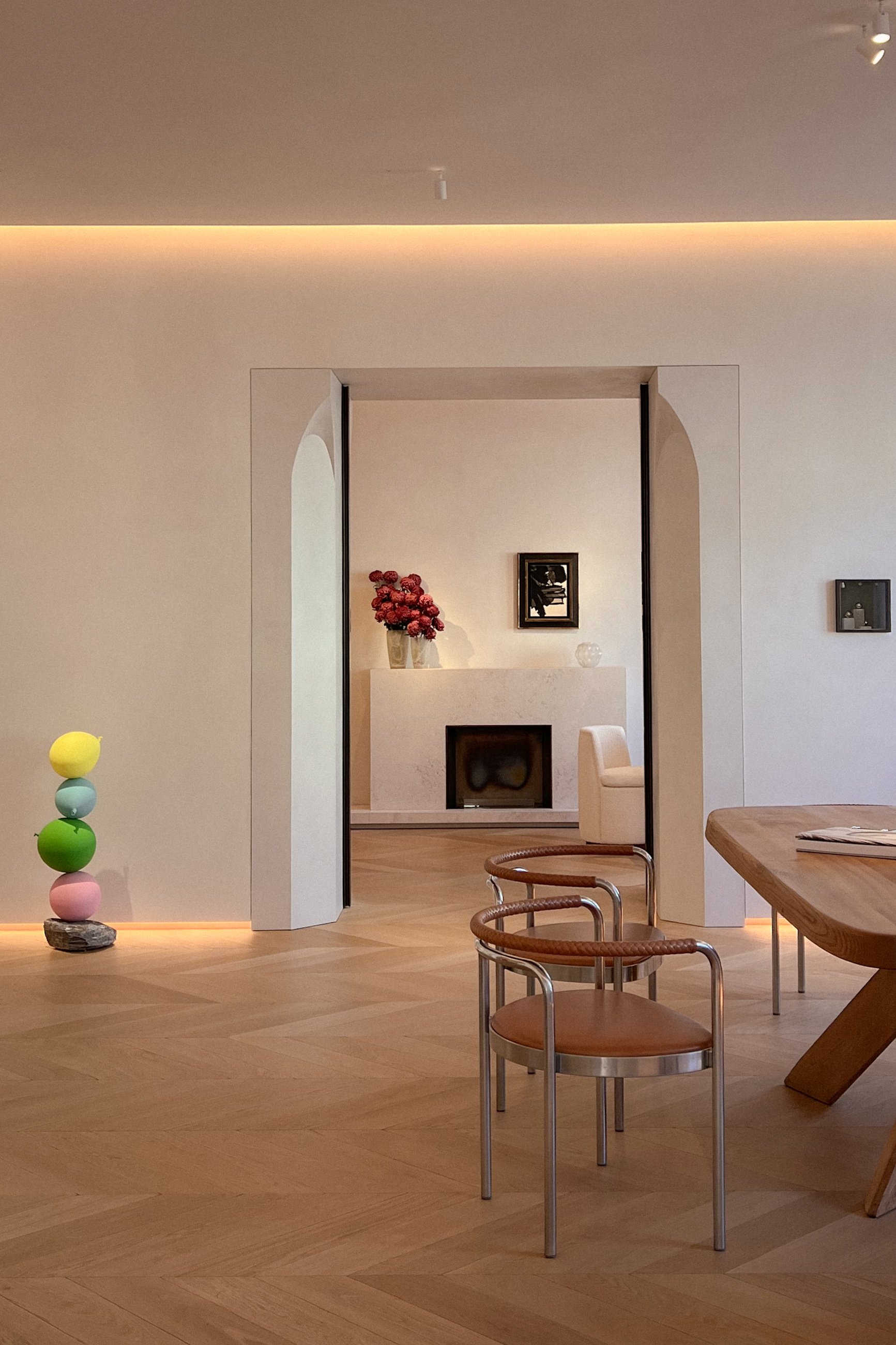 A modern, minimalistic interior featuring a light wood dining table, metal frame chairs with leather seats, a decorative sculpture with colored spheres on a rock, and a white fireplace adorned with a vase of flowers and artwork, with ambient lighting