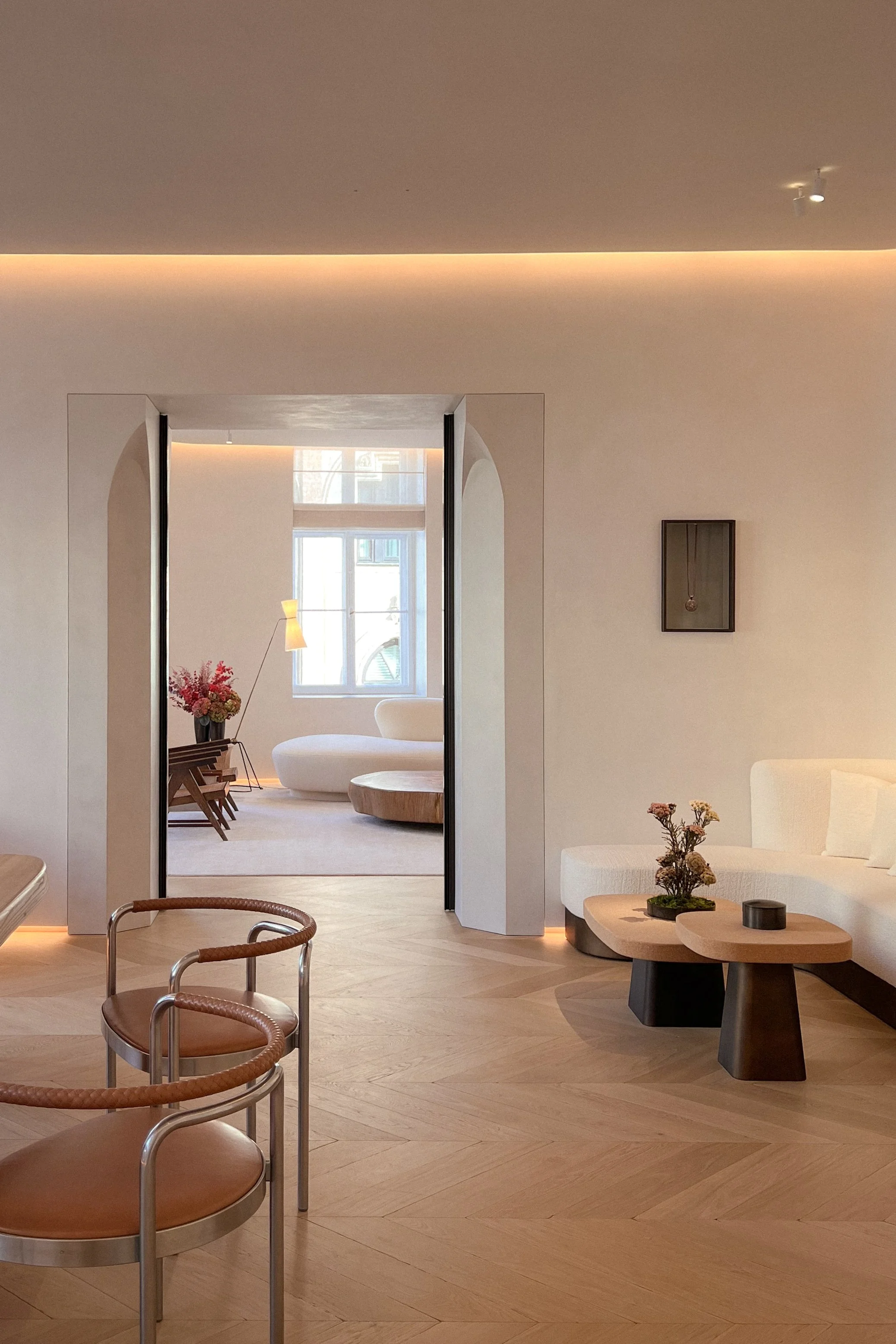 Modern living room with light-colored walls, wooden flooring, white sectional sofa, coffee tables, and decorative items. A doorway leads to another room with a large window, multi-level seating, and a flower arrangement.