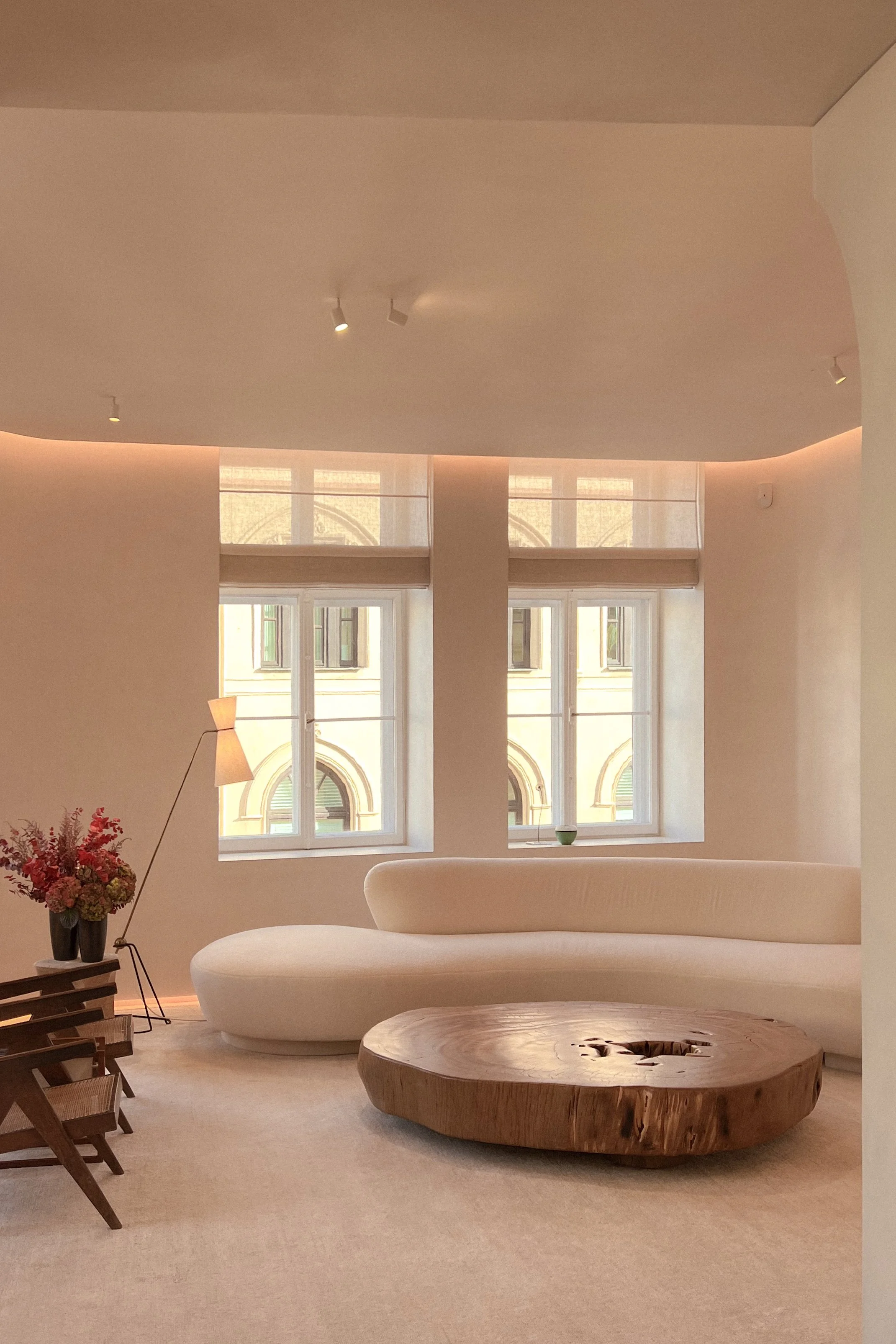 Minimalist living room with beige curved sofa, large wooden coffee table, flower vase, and window with beige blinds.
