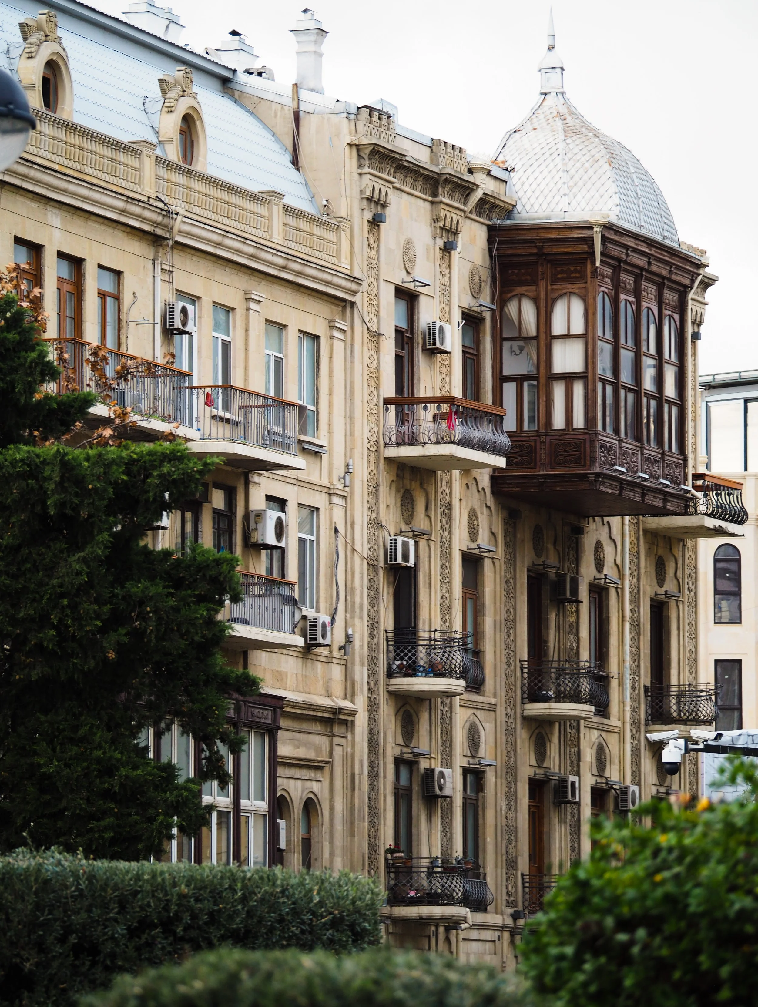 Ornate historical building with intricate balconies and a dome, surrounded by greenery, featuring classical architecture details and modern air conditioning units on facade.