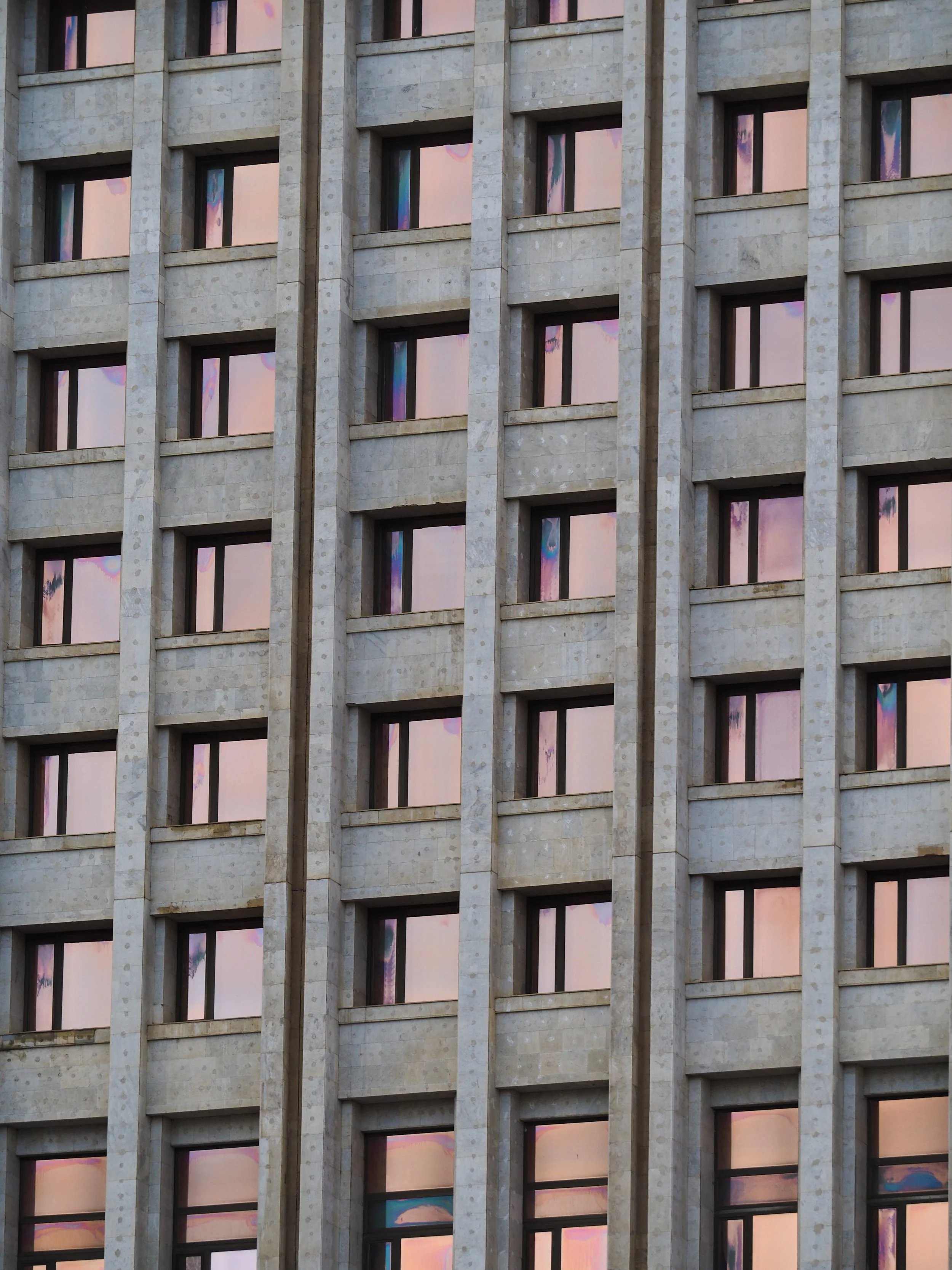 Exterior of a tall building with multiple windows reflecting a sunset, showcasing pink and purple hues.