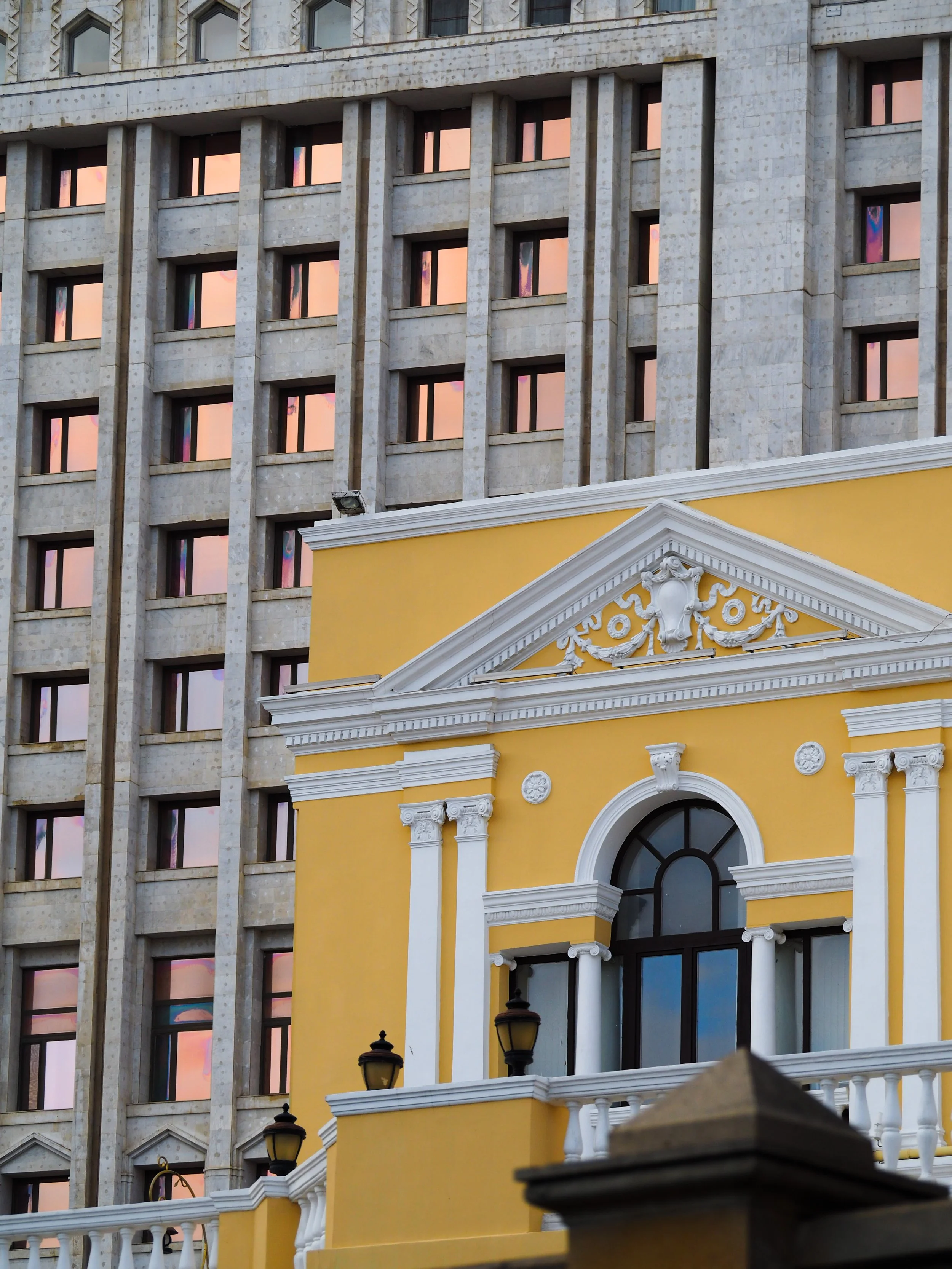 Facade of a yellow and white neoclassical building with decorative columns, an arched window, and architectural details, adjacent to a modern building with multiple windows reflecting a sunset hue.