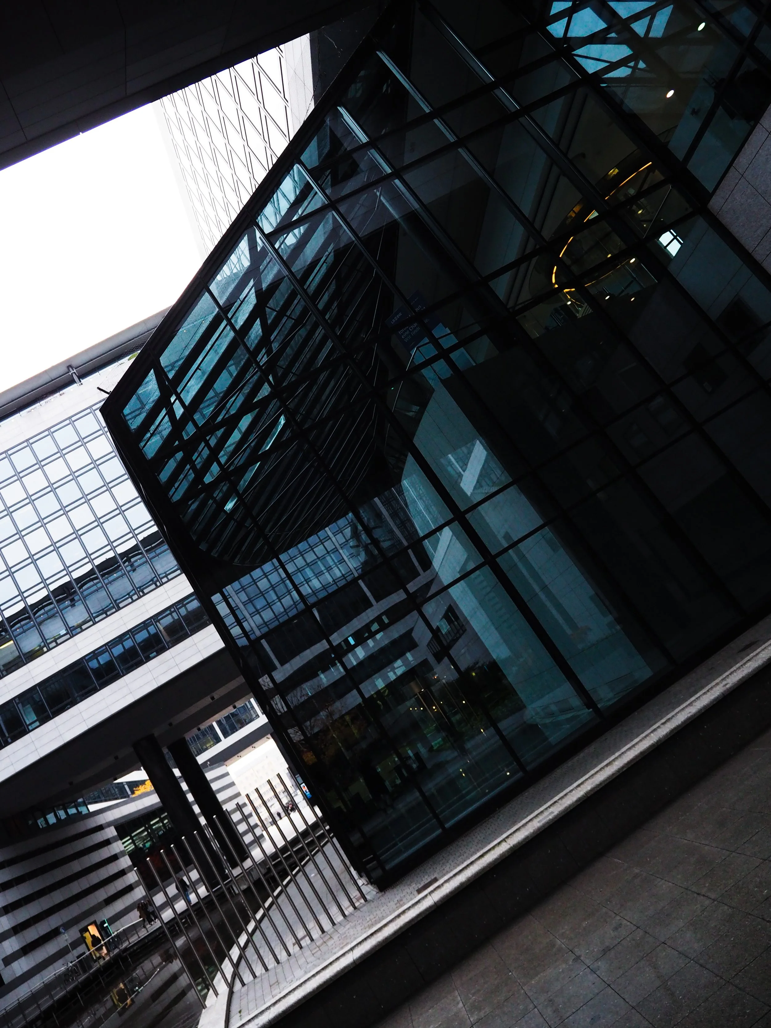 Modern glass and steel architectural building exterior with reflections, angular design, and surrounding structures in urban setting.
