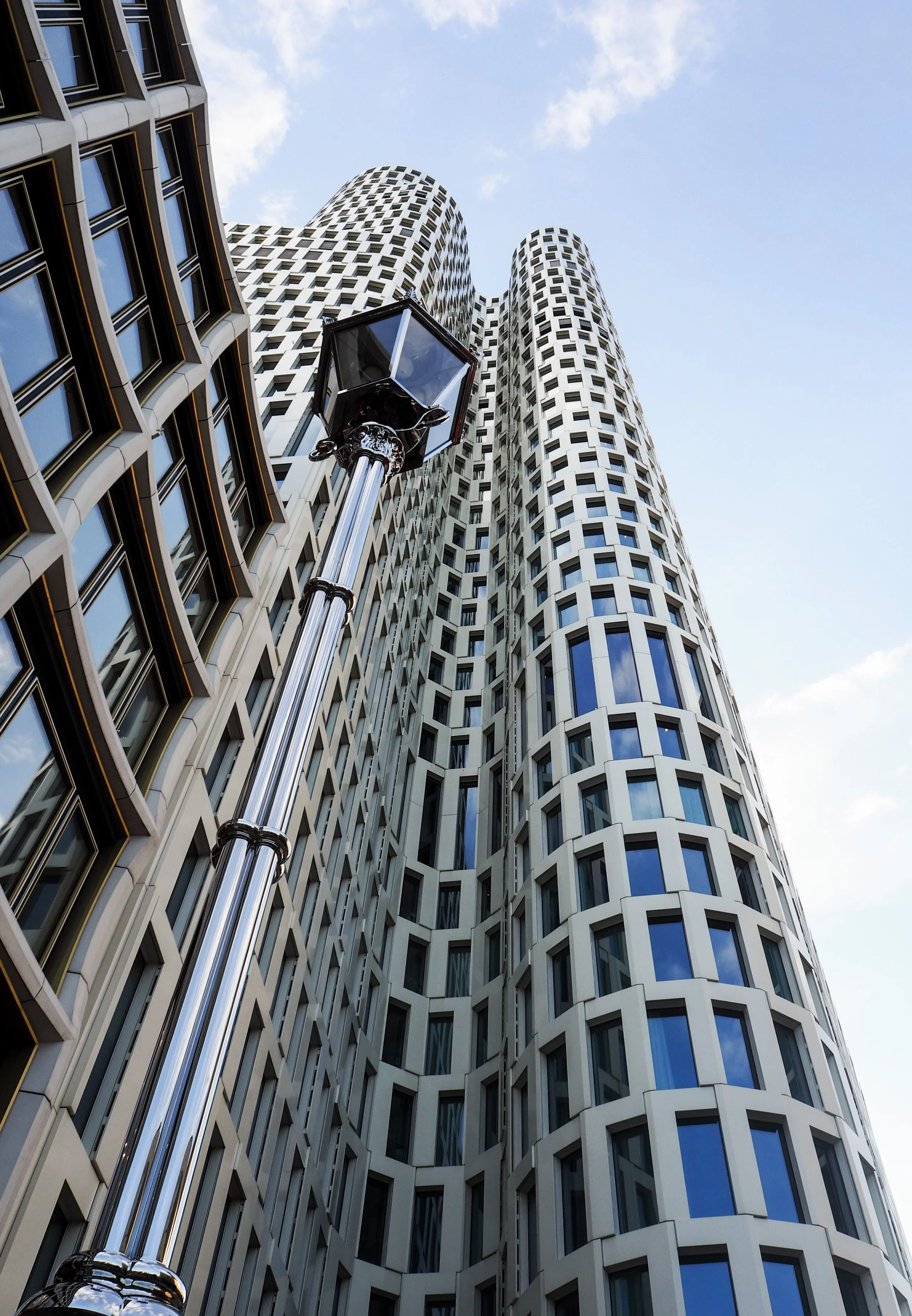 Modern high-rise building with a distinctive curved facade and large windows, viewed from below. A tall, shiny street lamp in the foreground.