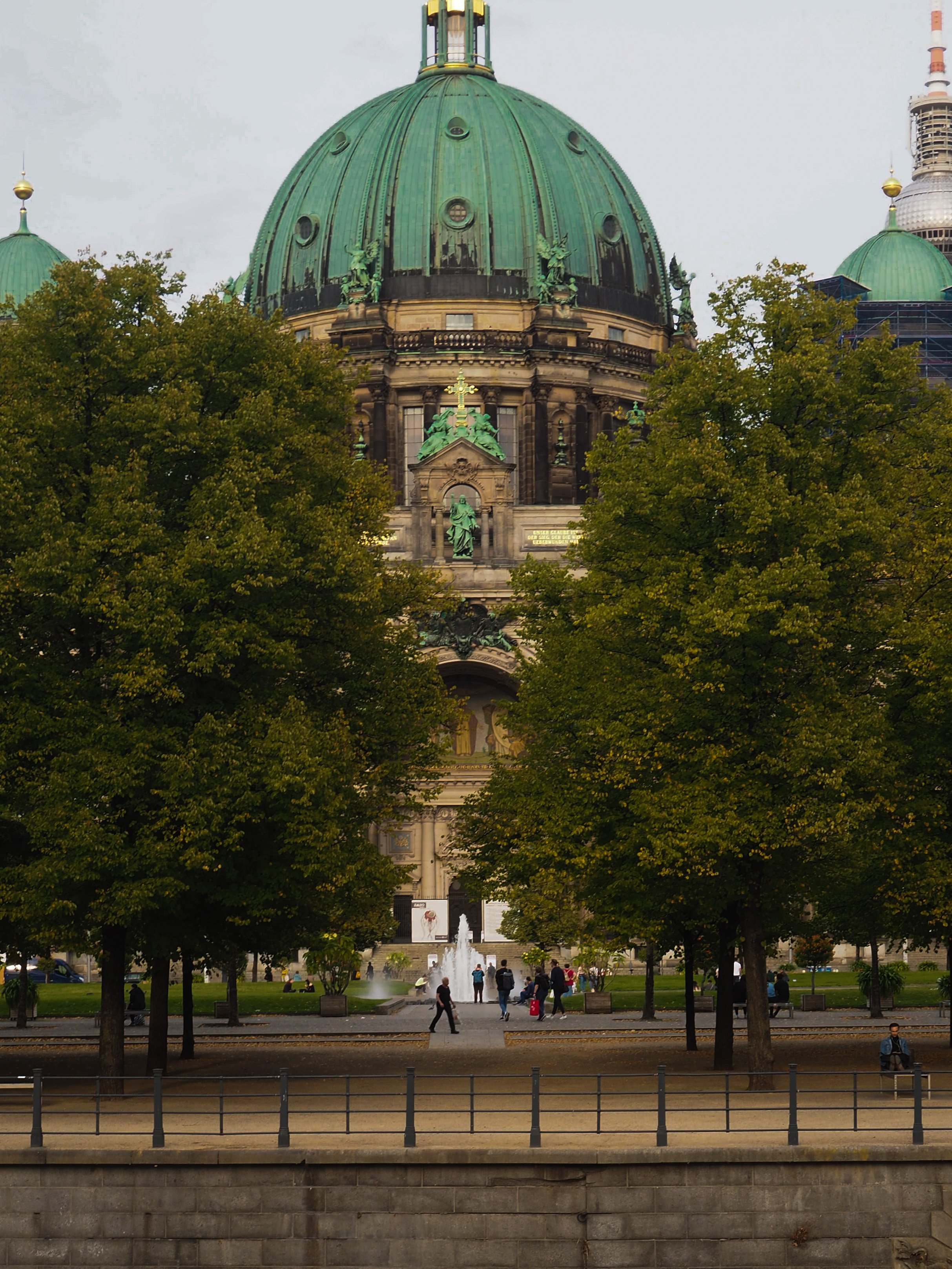 Large domed cathedral behind trees with people walking and sitting on benches, surrounded by fountains.