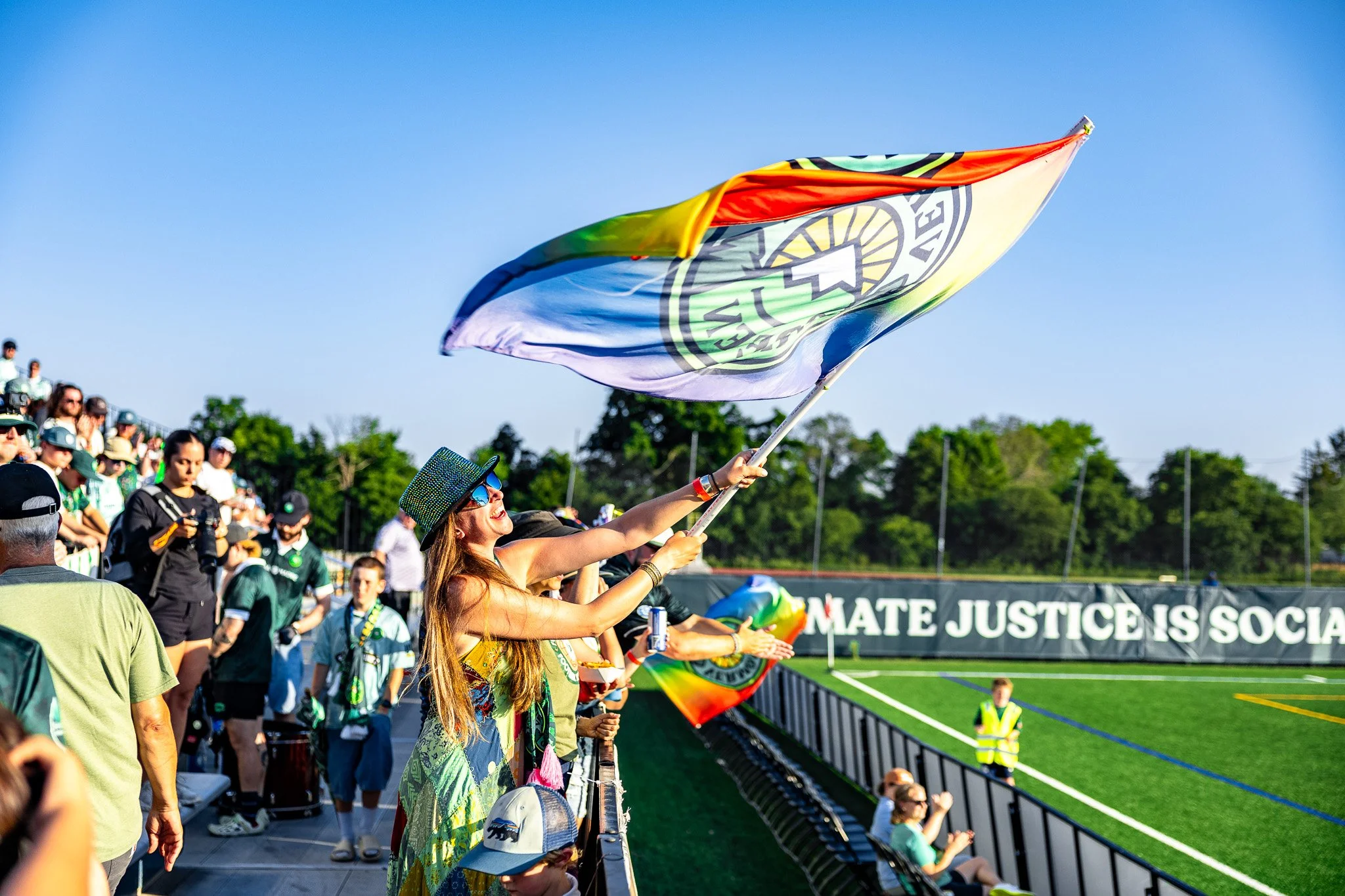 A woman waving a rainbow flag at a sports stadium during a peaceful event, with spectators in the background and a banner that reads 'Climate Justice is Social Justice'.